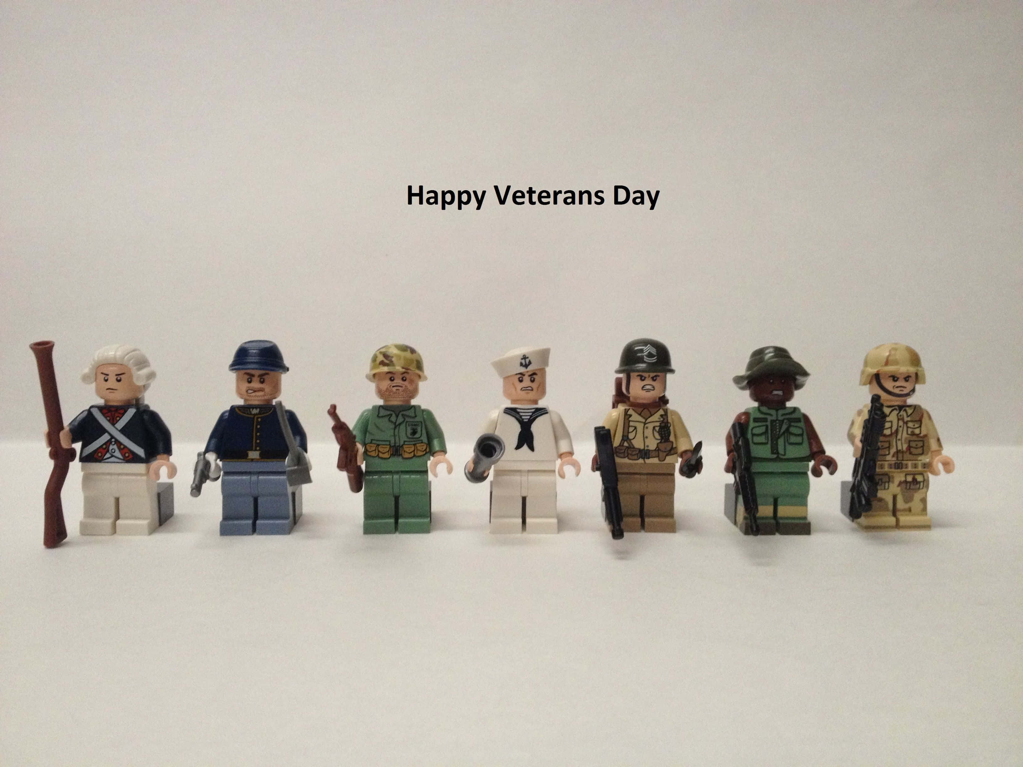 退伍軍人節 桌布 壁紙 — 七個穿著歷史軍服的LEGO士兵人偶在白色背景前排成一排，上方有「Happy Veterans Day」文字。