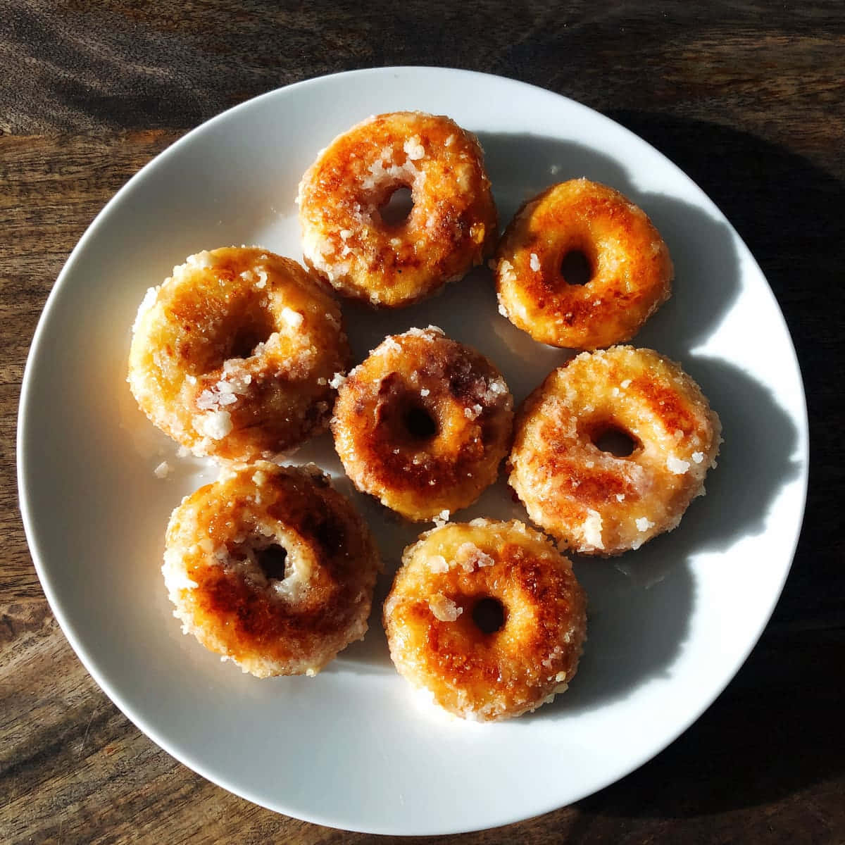 Glazed Donut 桌布 壁紙 — 七個金黃色糖霜甜甜圈放在深色木桌上的白色碟子上。