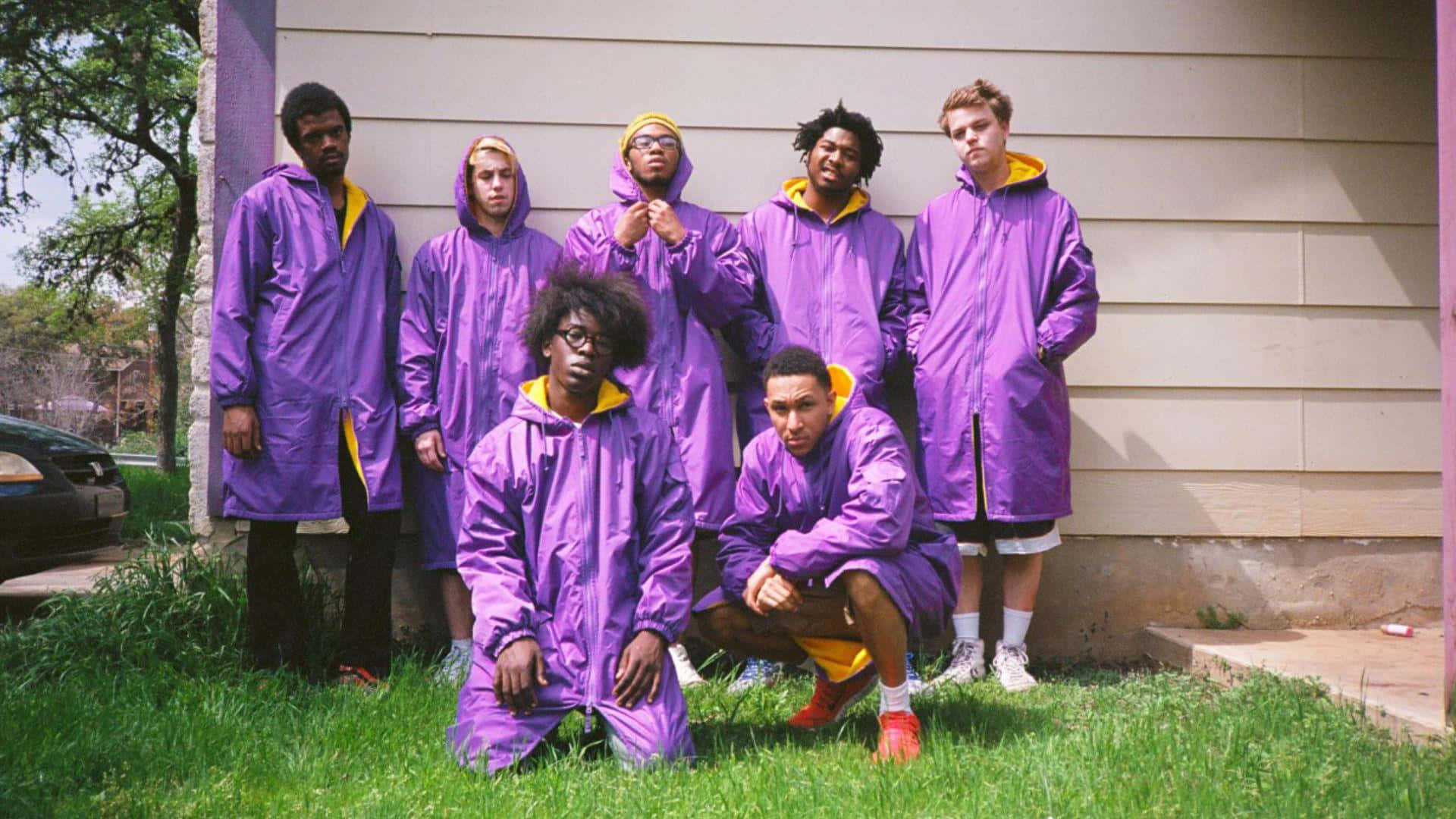 Brockhampton 桌布 壁紙 — 七名年輕男子身穿相配的紫色雨衣和黃色連帽，在米色房屋前的草地上自信地擺姿勢。