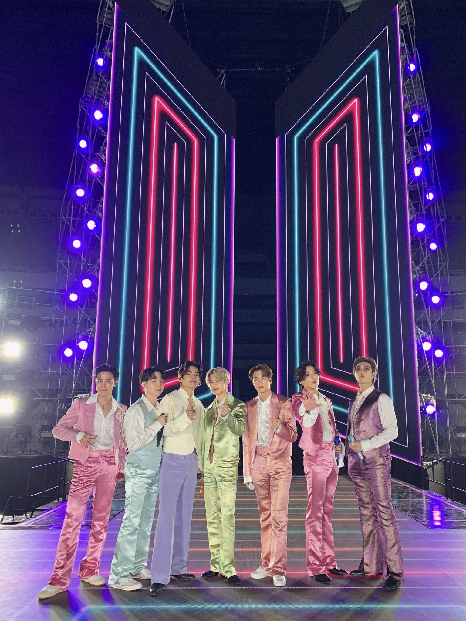 bts group cute 桌布 壁紙 — 七名身穿彩色緞面西裝的男子在霓虹燈舞台上自信擺姿勢，舞台由發光的粉紅色和藍色幾何面板框住。