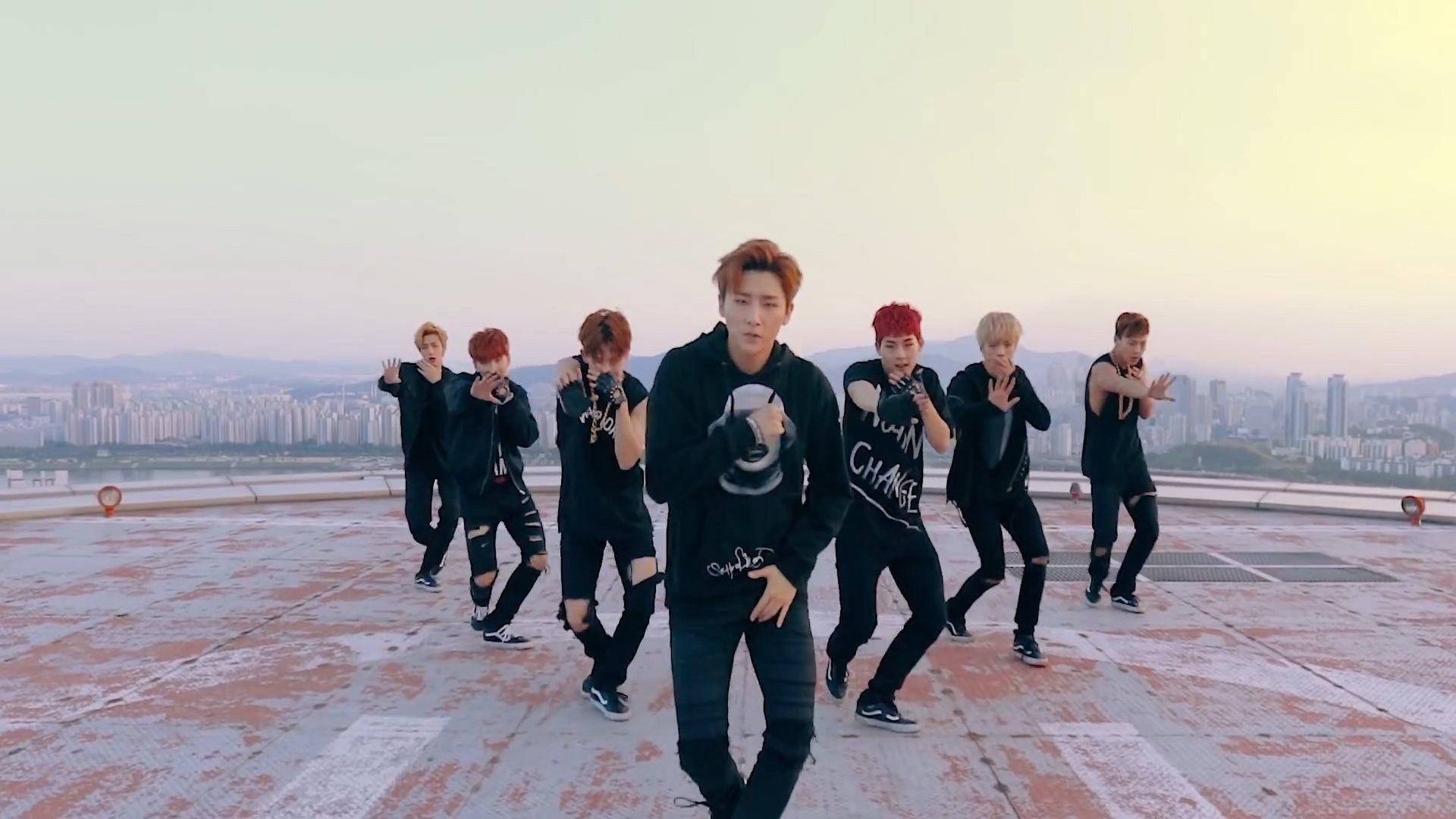 Monsta X Hero 壁紙 — 七名身穿黑色服裝的男舞者在一個風化的屋頂上表演同步編舞，背後是廣闊的城市景觀和朦朧的日落。