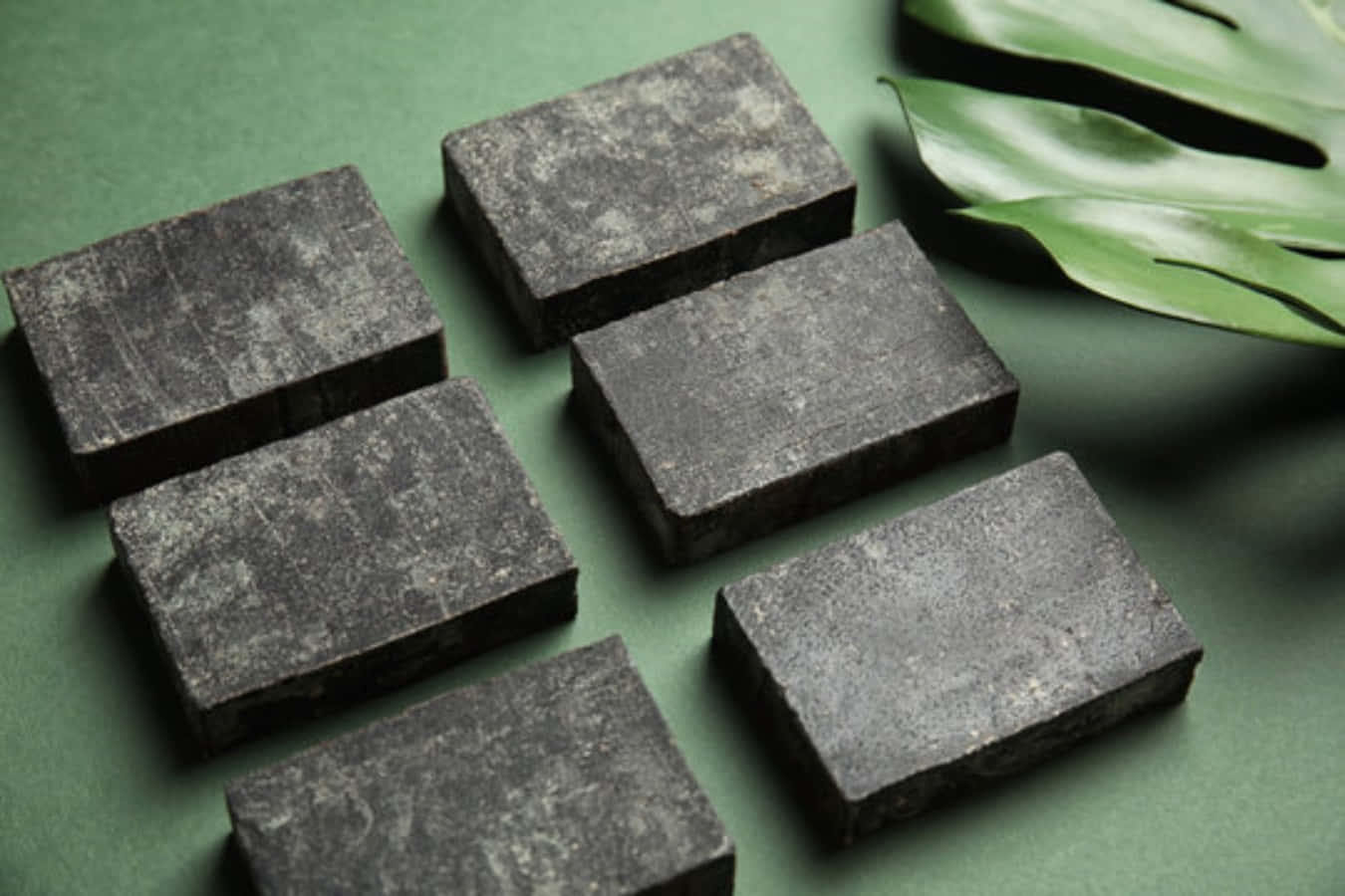 Black Soap 桌布 壁紙 — 七塊深色紋理長方形塊排列在柔和的綠色表面上，旁邊有一塊大的光澤龜背竹葉。