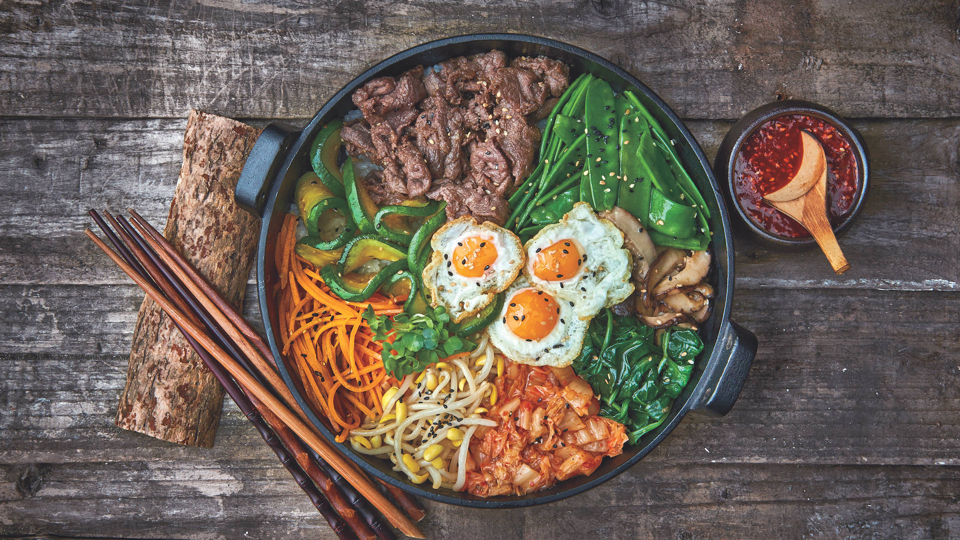 三人份誘人韓式拌飯盛宴 壁紙 — 一碗色彩繽紛的韓國石鍋拌飯，配有牛肉、雞蛋、蔬菜和泡菜，放在質樸的木桌上，附有筷子和辣椒醬。
