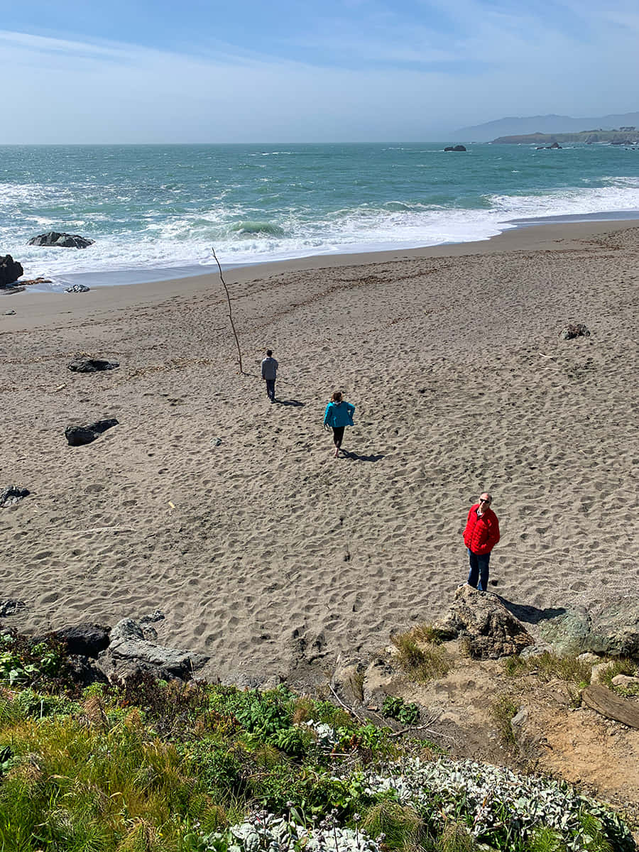 Portuguese Beach 桌布 壁紙 — 三人在陽光明媚的沙灘上漫步，碧綠的海浪拍打著岩石，天空湛藍清澈。