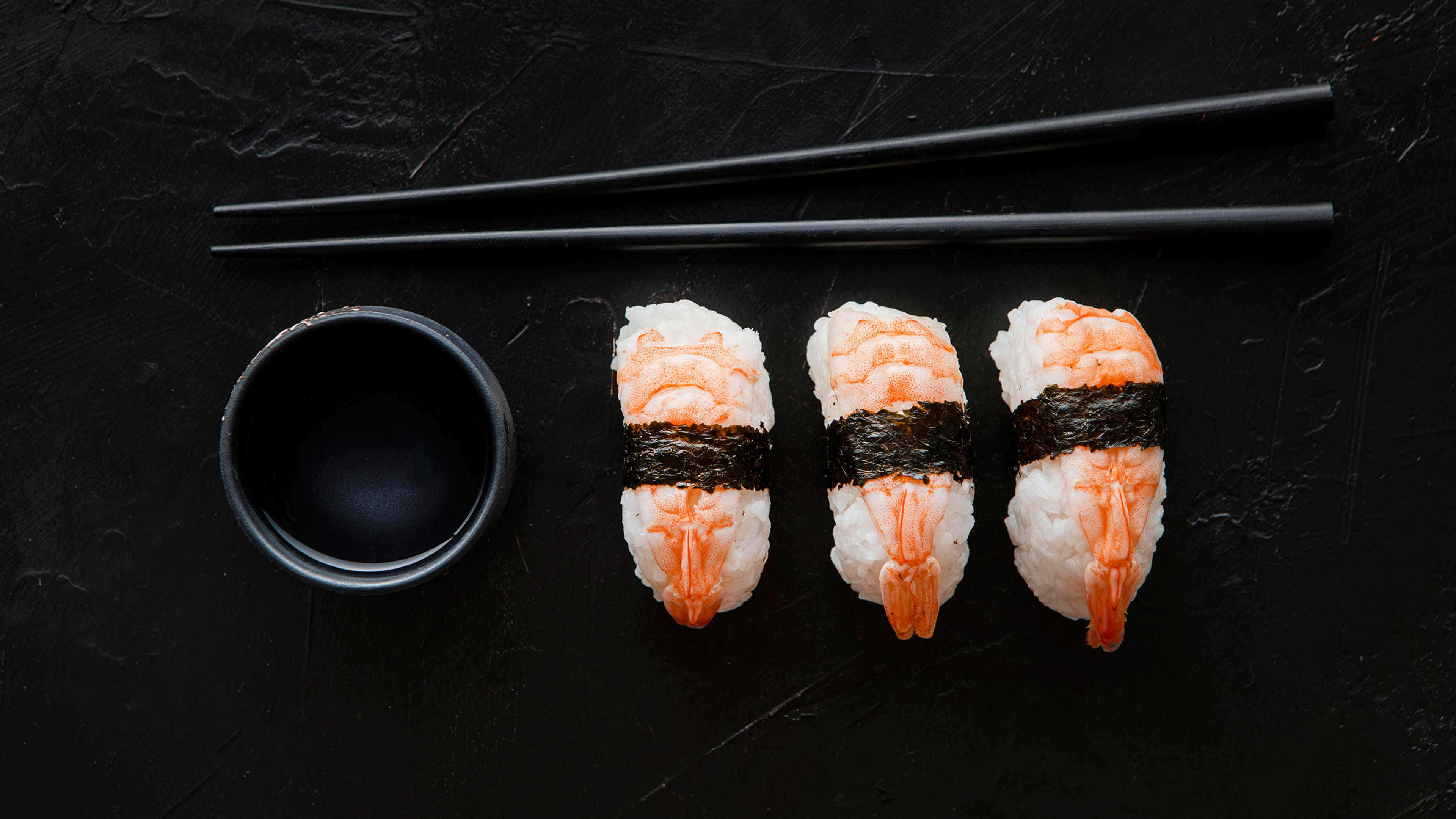 Sushi 桌布 壁紙 — 三件帶有黑色海苔帶的蝦握壽司整齊地放在一個小碗和黑色筷子旁邊，置於有紋理的深色石板表面上。