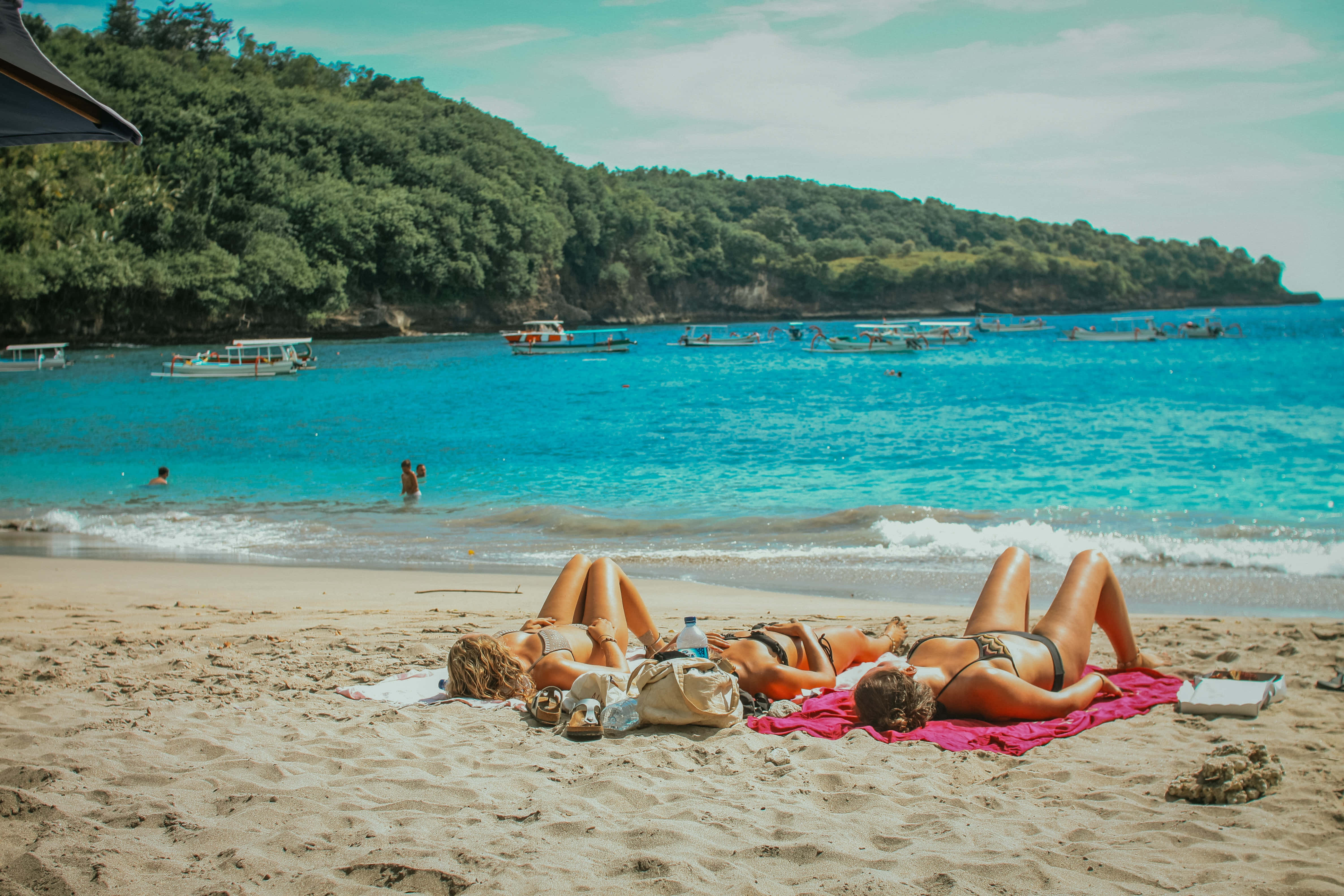 Spanish Beach 桌布 壁紙 — 三位女士在明亮的天空下，在沙灘上放鬆，背景是碧綠的海水和樹木繁茂的山丘。
