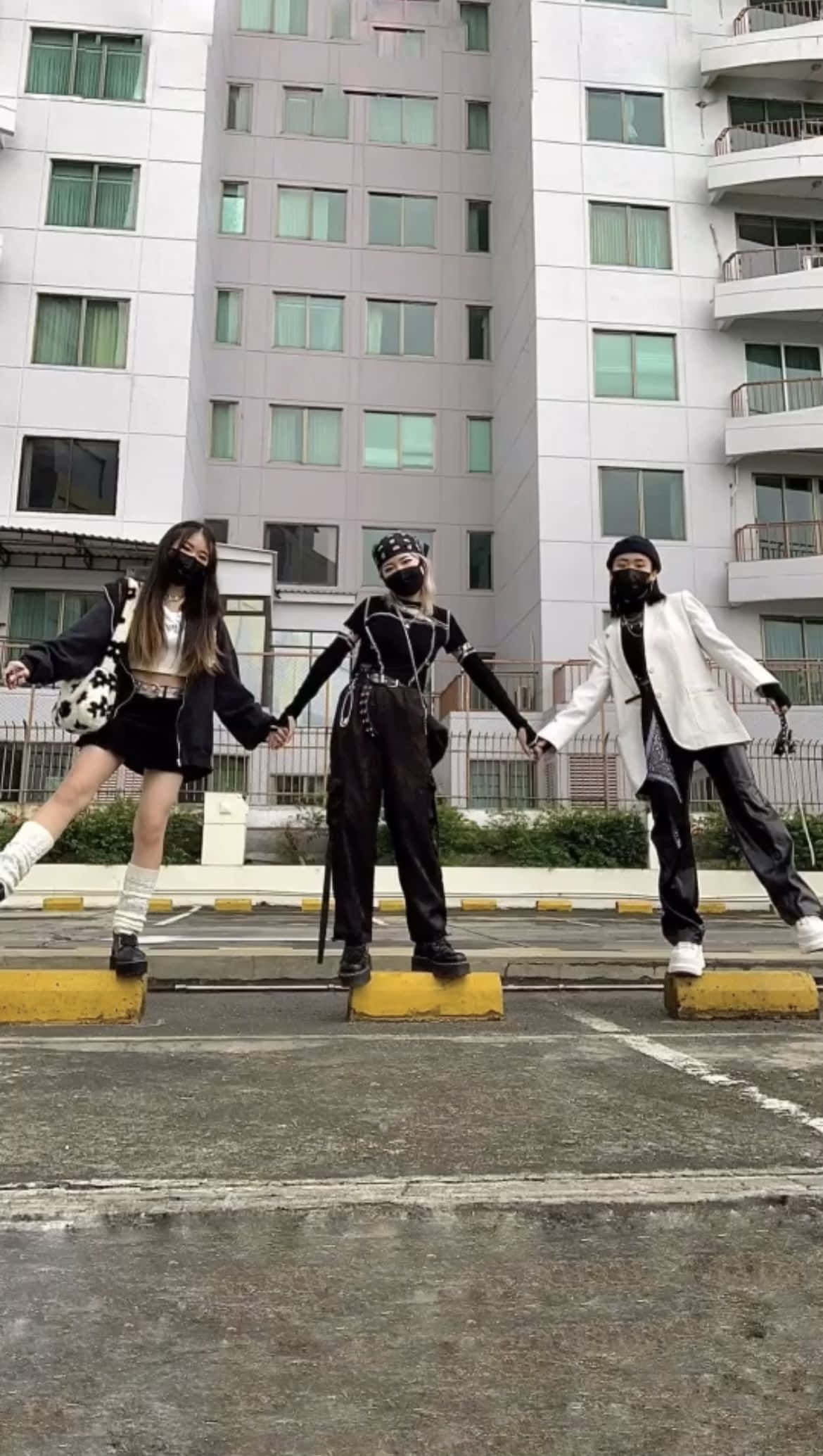 三位好友停車場合影 壁紙 — 三位戴著口罩的年輕女性穿著前衛街頭服飾，在現代公寓大樓前的黃色停車塊上擺出同步姿勢。