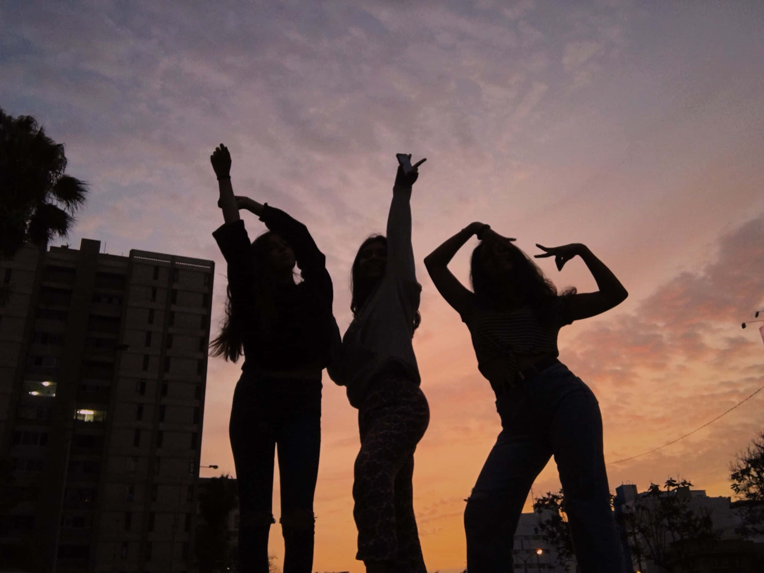 三位好友夕陽剪影 壁紙 — 三位剪影朋友在充滿活力的日落天空背景下，以城市建築為背景擺出俏皮的舞蹈姿勢。