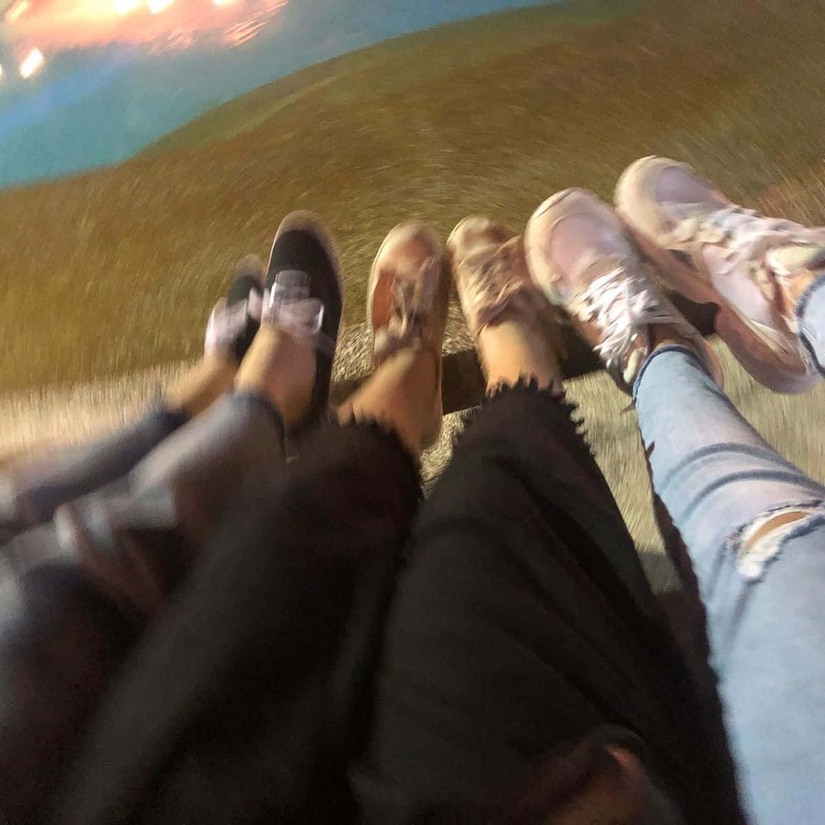 三位好友的鞋履特寫 壁紙 — 四個人的腳穿著不同的鞋子和牛仔褲，一起站在草地上，頭頂是色彩繽紛的暮色天空。