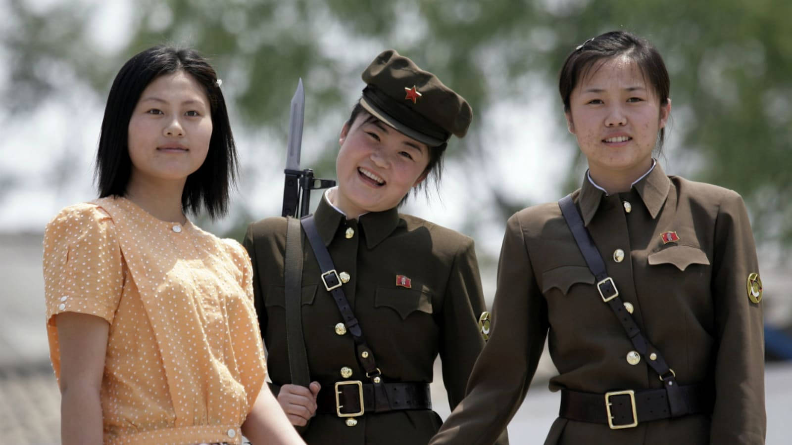 North Korea 桌布 壁紙 — 三位年輕女性站在一起，一位身穿軍服手持步槍，另外兩位微笑，其中一位穿著波點裙。