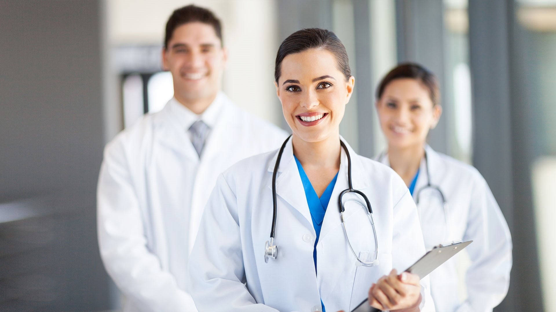 Stethoscope 桌布 壁紙 — 三位微笑的醫護專業人士身穿白袍，站在明亮現代化的醫院走廊，其中一人拿著記事板。