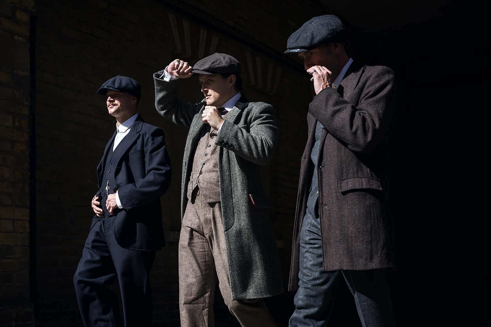 Peaky Blinders 桌布 壁紙 — 三位戴著復古平頂帽、身穿西裝的男士，在戲劇性的陽光下靠著磚牆站立，擺出自信的表情。