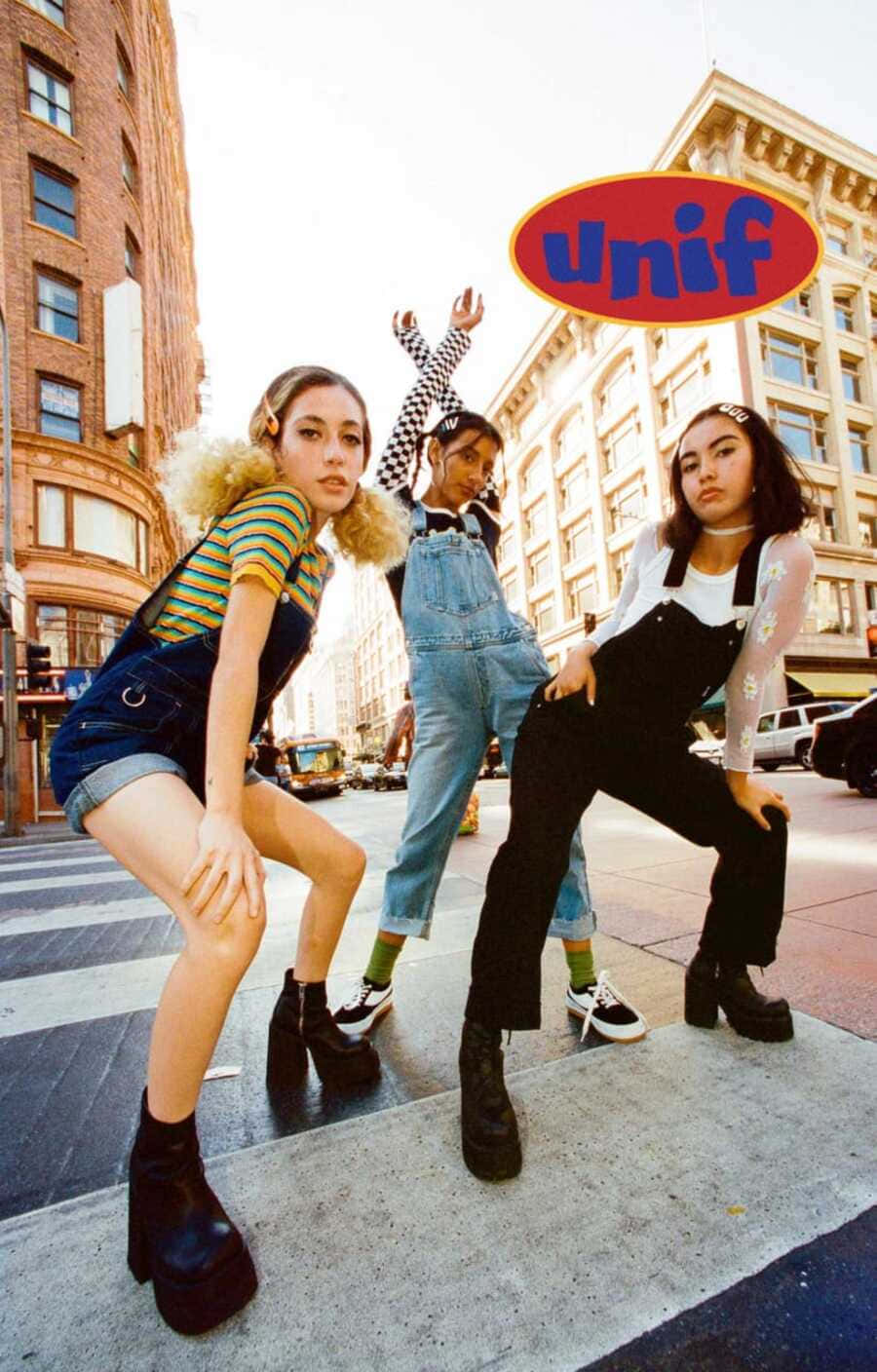 Baddie Vintage 90's Aesthetic 桌布 壁紙 — 三位時尚年輕女性在明亮的日光下於城市街道上擺出大膽姿勢，上方漂浮著色彩繽紛的「unif」標誌。