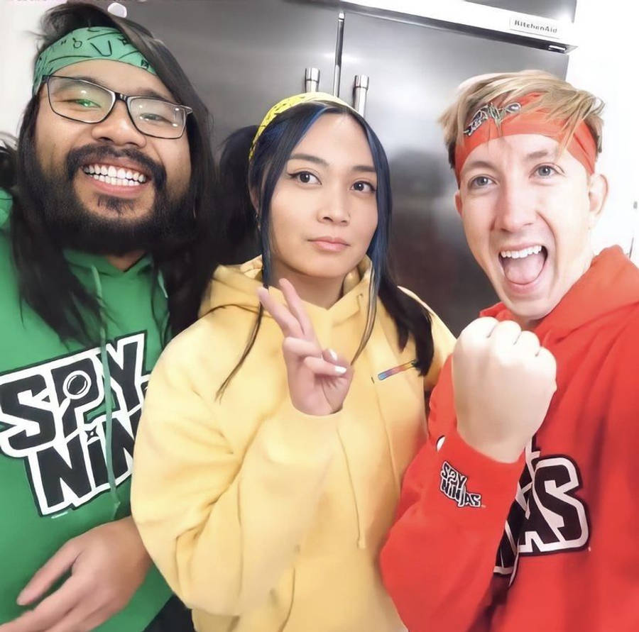 微笑 桌布 壁紙 — 三位朋友穿著色彩繽紛的Spy Ninjas連帽衫，在身後有不銹鋼雪櫃的廚房裡愉快地擺姿勢。