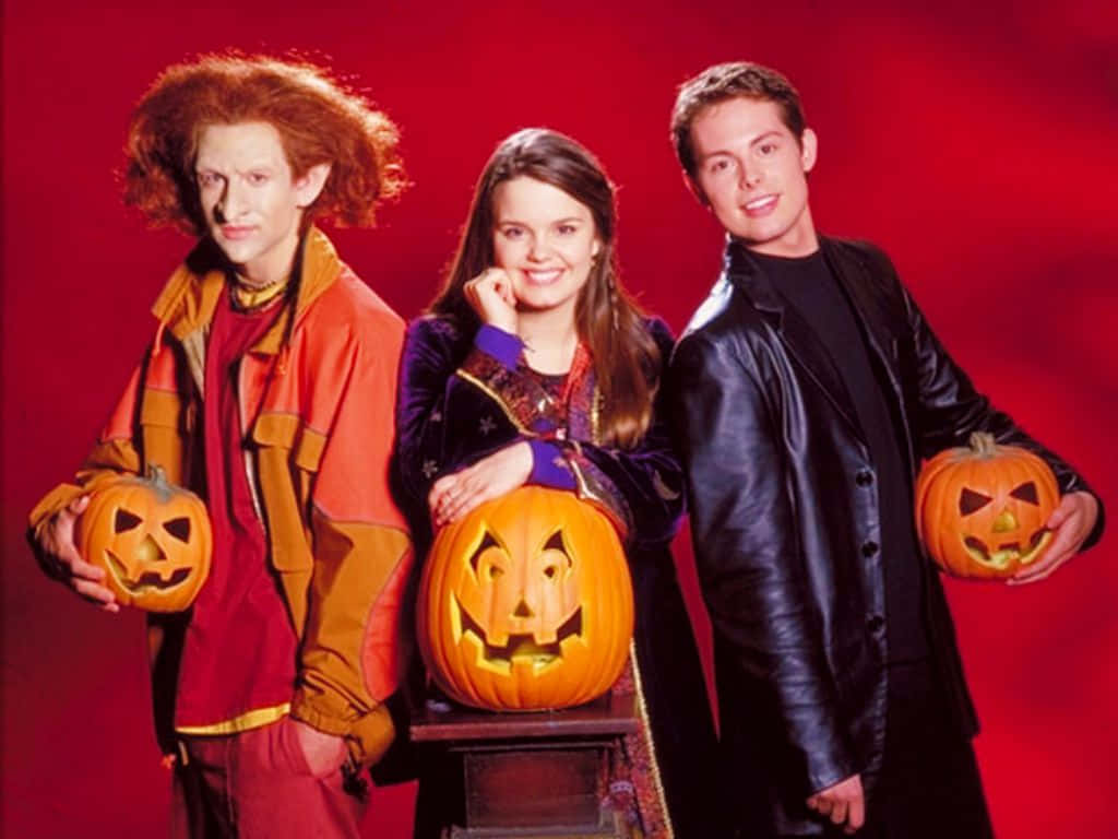 Halloweentown 桌布 壁紙 — 三位演員穿著萬聖節服裝，在鮮紅色的攝影棚背景前，手持雕刻南瓜擺姿勢。