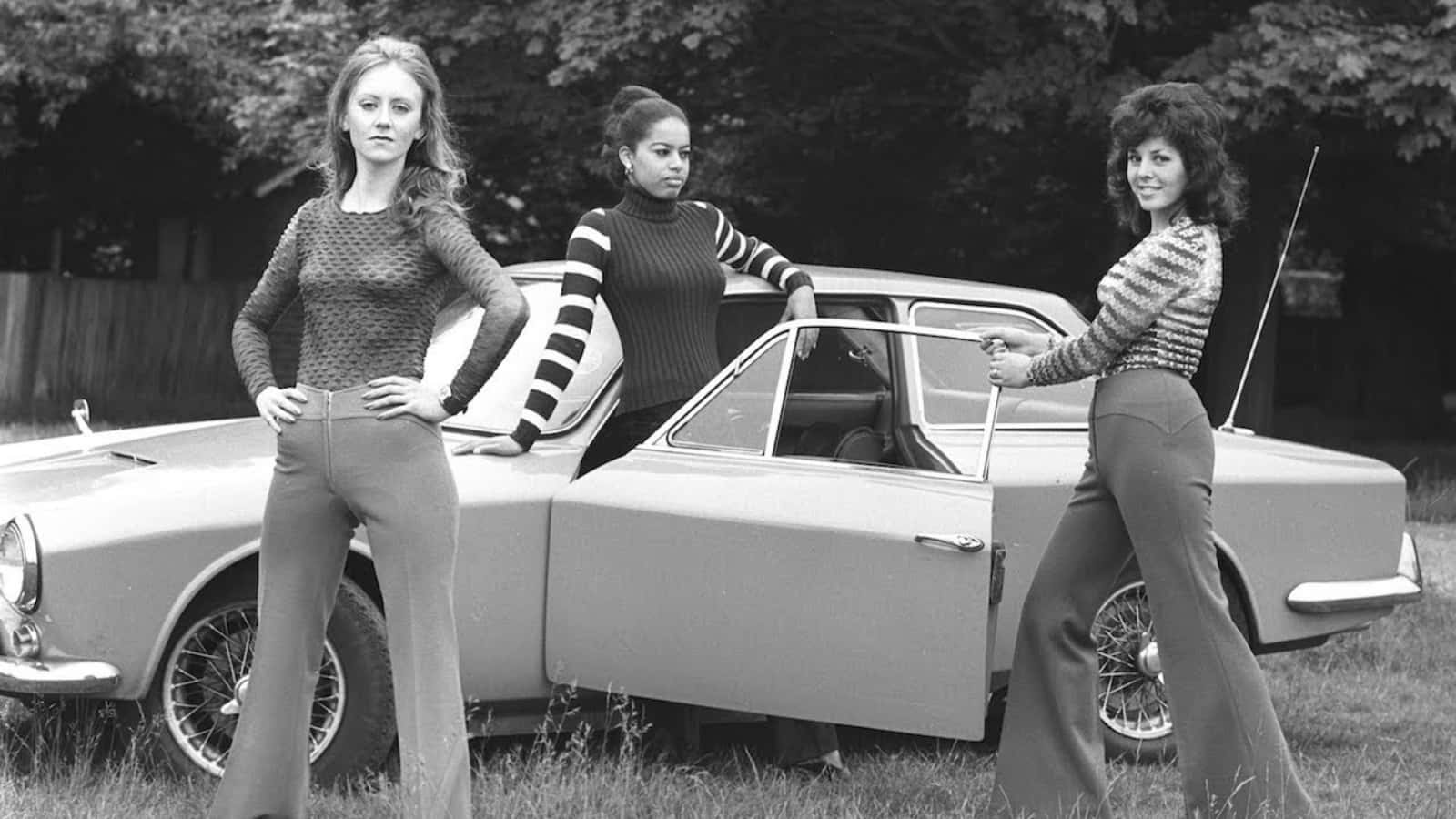三位穿著1970年代服裝的女性，在草地上站在一輛復古汽車旁。 — 70年代gif 壁紙