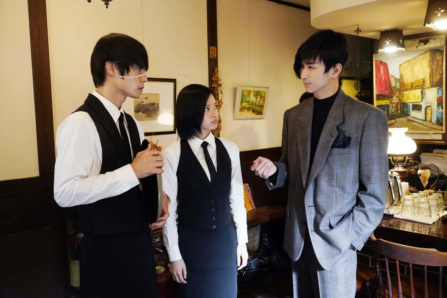 Tokyo Ghoul Movie 桌布 壁紙 — 三位穿著正裝的年輕人站在一間燈光溫暖、擺設木製家具和牆上掛有裱框畫作的復古咖啡館內。