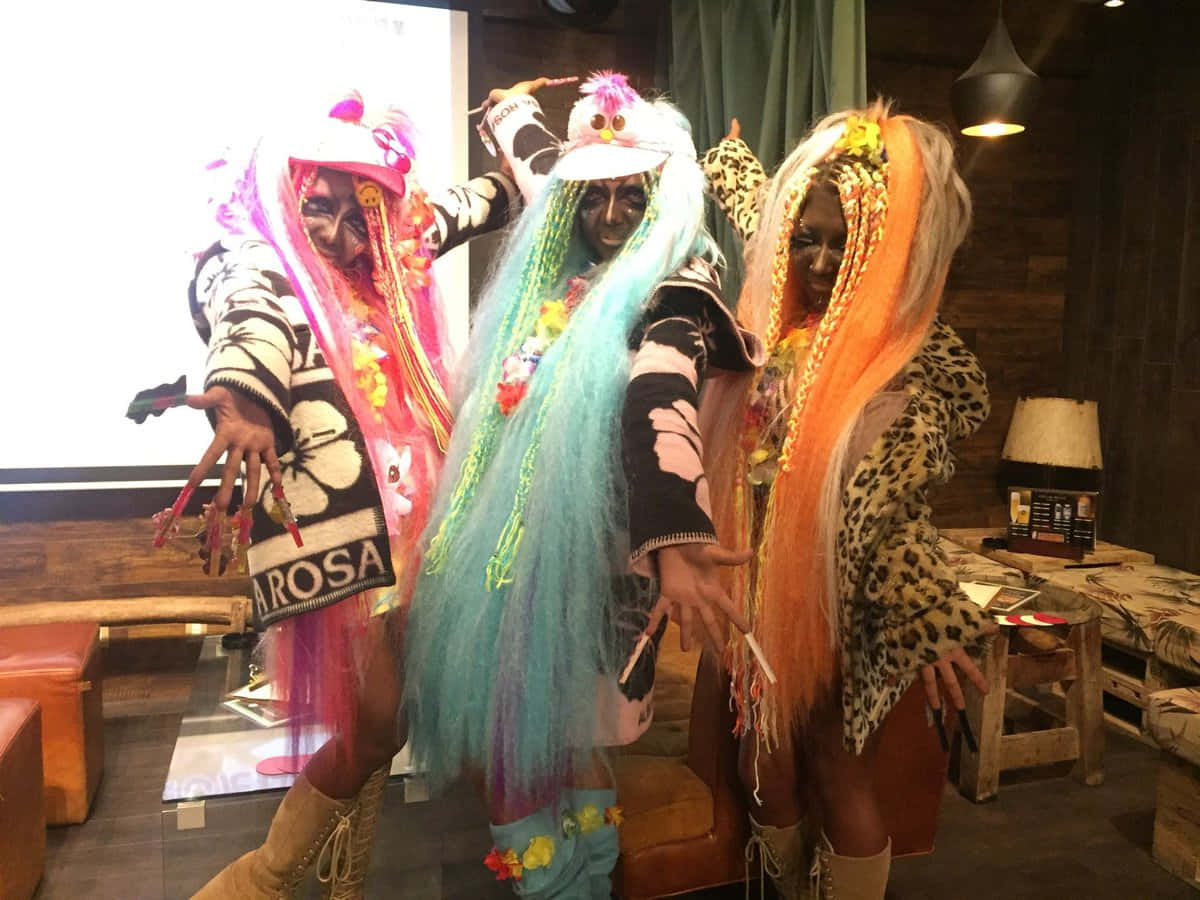 Gyaru Fashion 桌布 壁紙 — 三位穿著精緻服裝、戴著鮮豔彩色假髮、化著大膽妝容的表演者，在質樸的室內休息室中戲劇性地擺出姿勢。