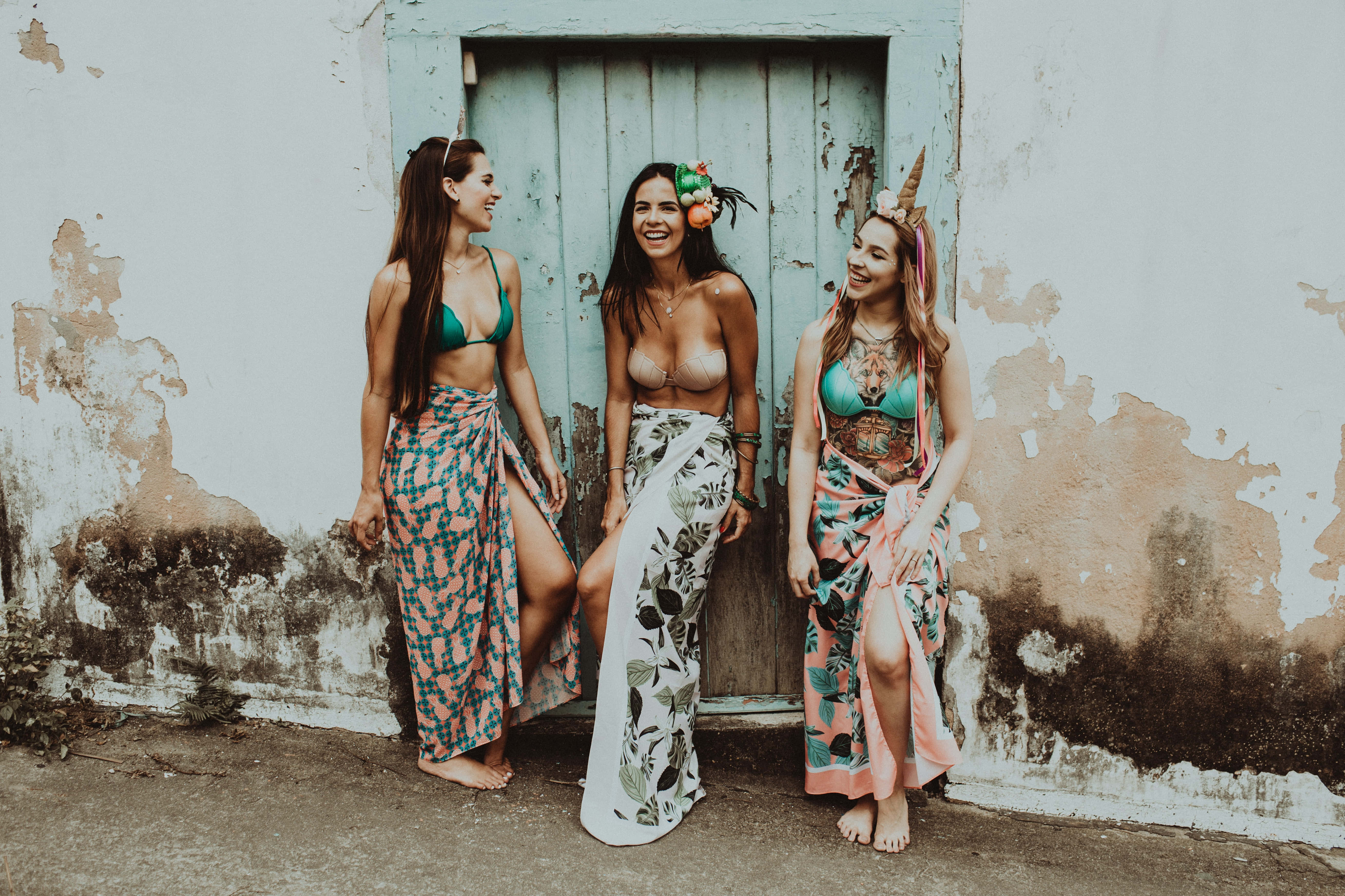 Summer Fashion 桌布 壁紙 — 三位笑容滿面的女士身穿色彩繽紛圖案沙龍和比堅尼，在鄉村風格的牆上，靠著一扇褪色的藍色門，俏皮地擺姿勢。