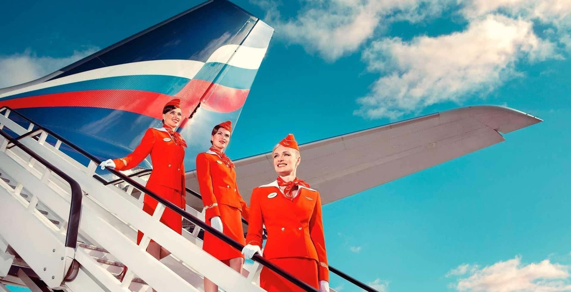 Flight Attendant 桌布 壁紙 — 三位笑容滿面的空中服務員身穿鮮紅色制服，站在飛機樓梯上，背景是晴朗的藍天和蓬鬆的白雲。