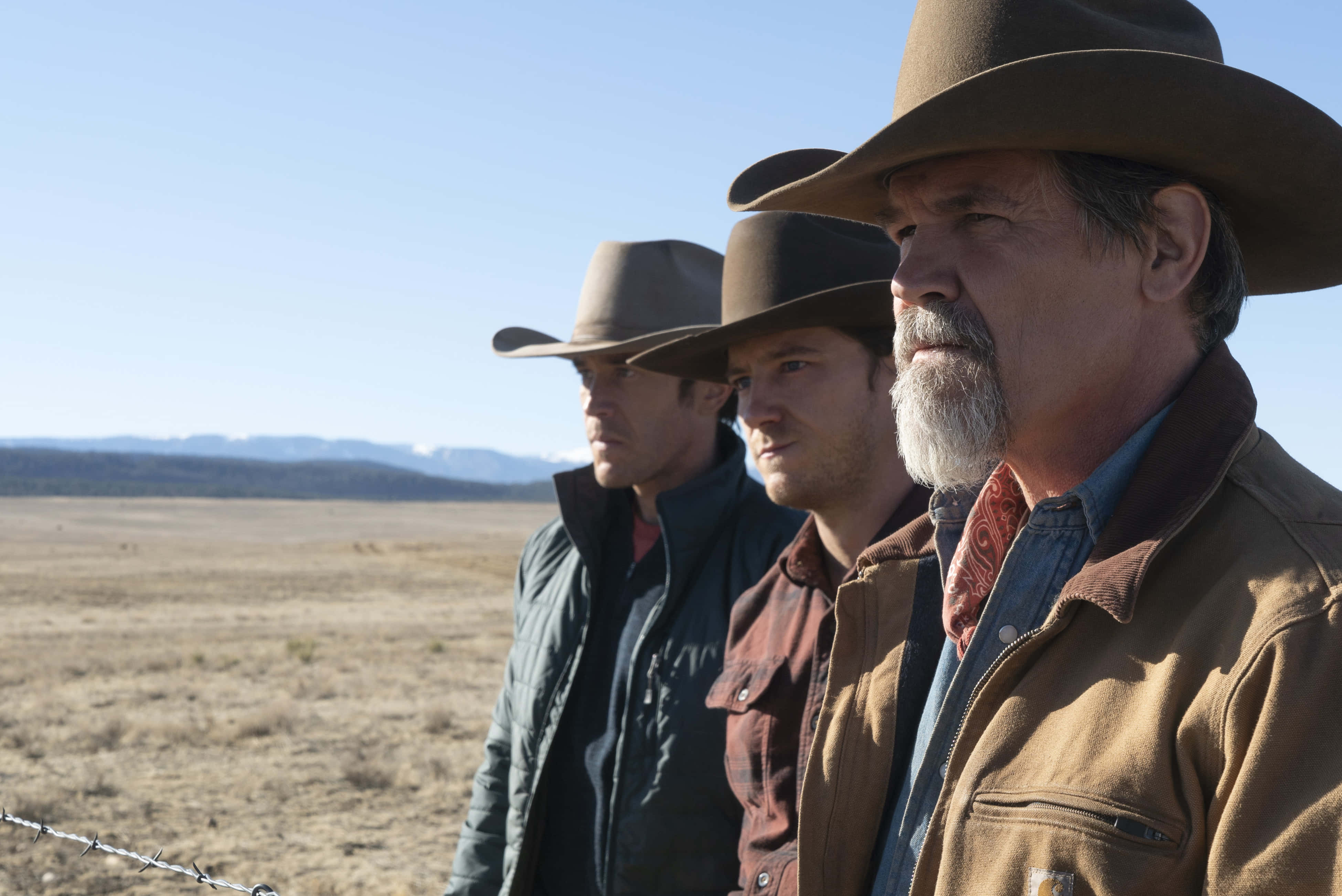 Josh Brolin 桌布 壁紙 — 三位粗獷的牛仔戴著寬邊帽，站在陽光明媚的沙漠平原上，遠山在清澈的藍天下。