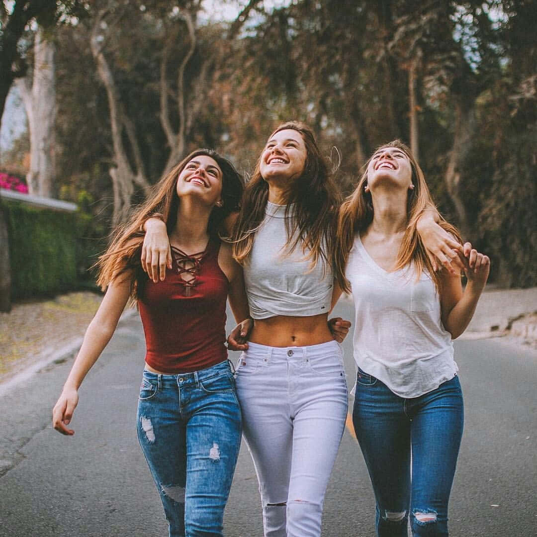 三位美女同行圖 壁紙 — 三位快樂的年輕女性在陽光明媚的日子裡，笑著手挽手走在綠樹成蔭的街道上。