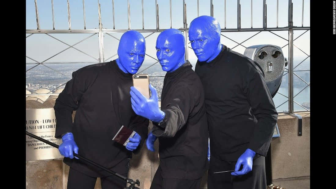Blue Man Group 桌布 壁紙 — 三位藍面表演者身穿黑色服裝，在高聳的觀景台上自拍，俯瞰廣闊的城市景觀。