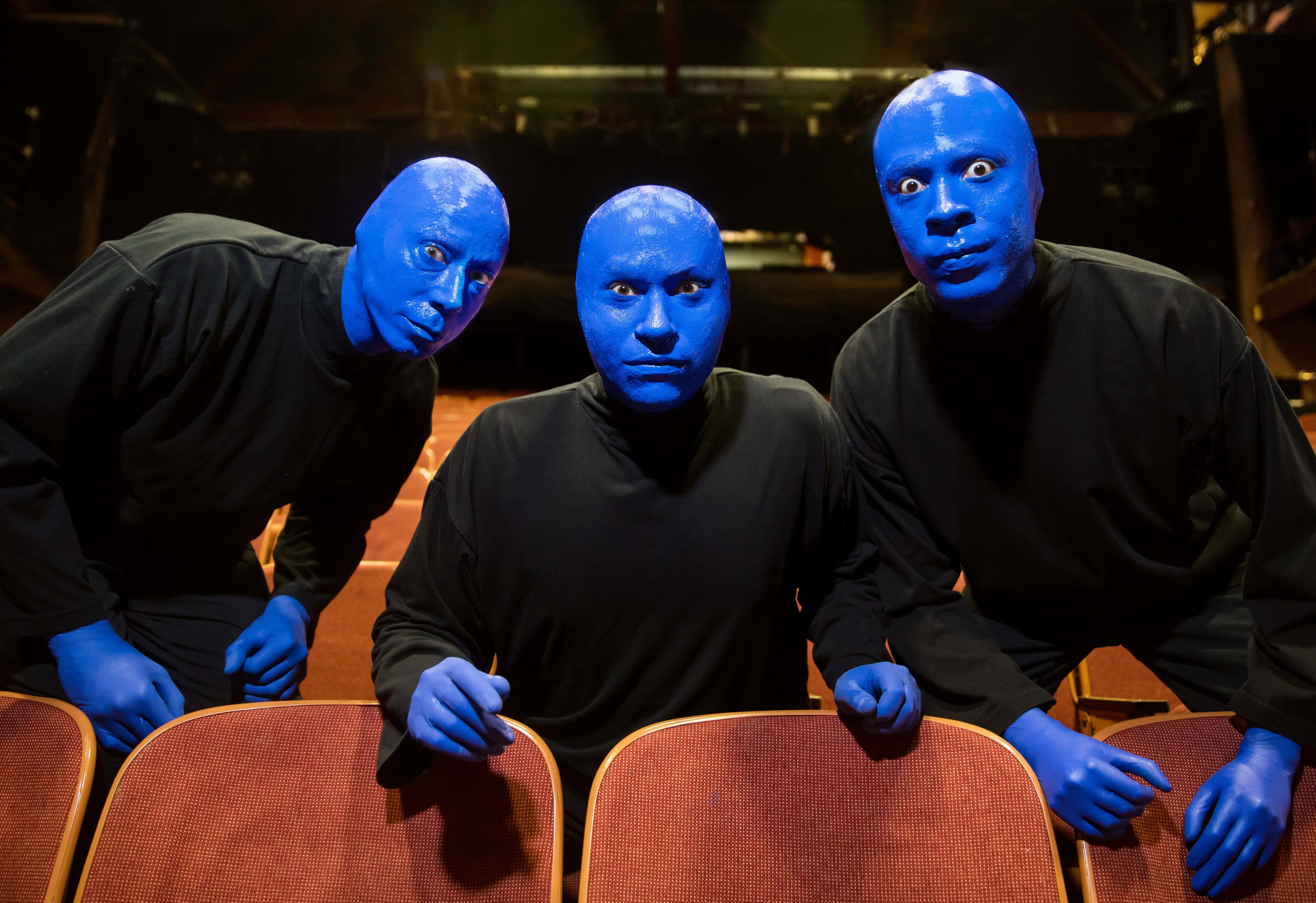 Blue Man Group 桌布 壁紙 — 三位表演者臉上和手上塗滿藍色顏料，在昏暗的舞台場景中俯身靠在紅色劇院座位上。