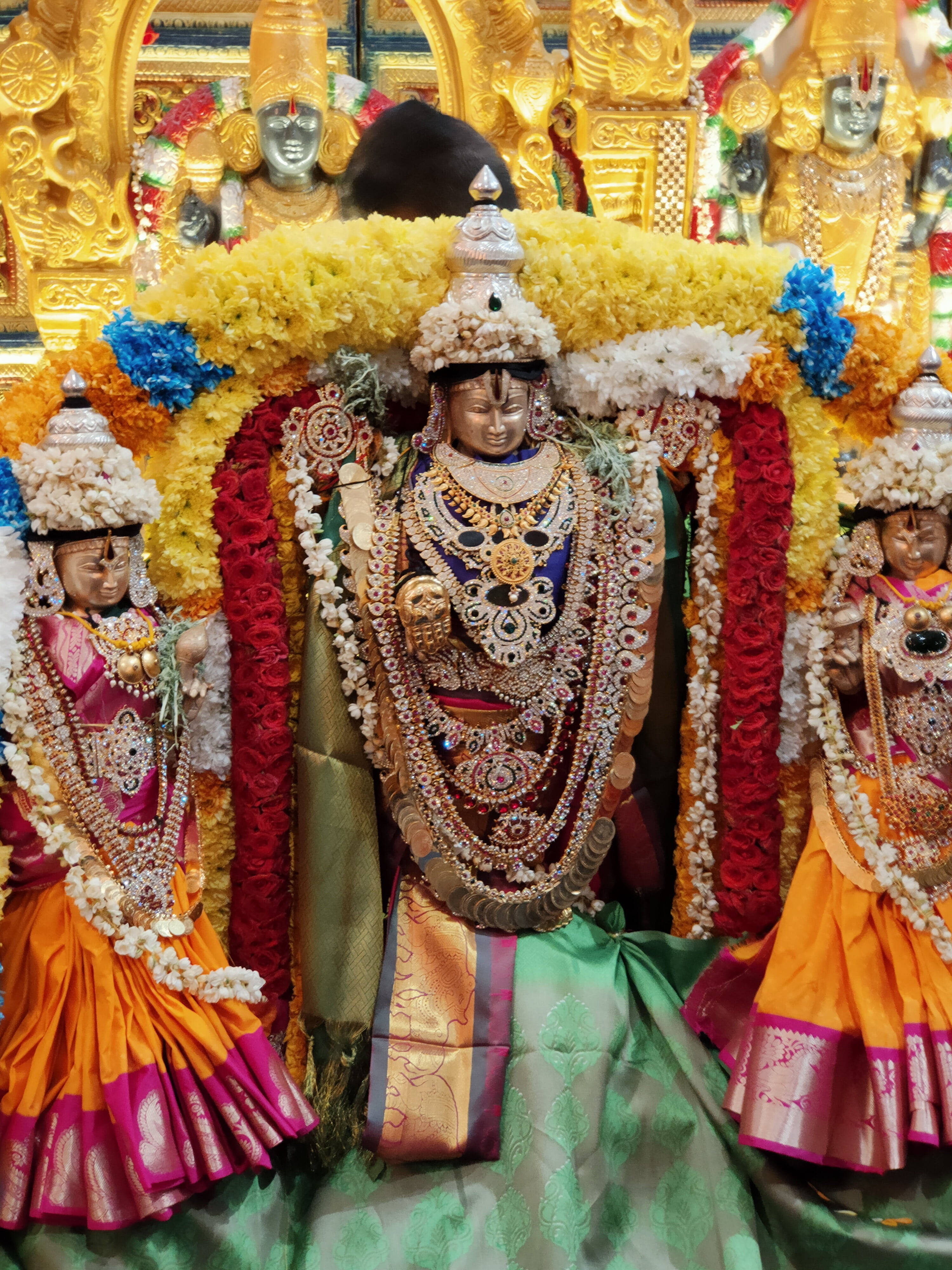 Lord Balaji 桌布 壁紙 — 三位裝飾華麗的印度教神祇，佩戴金飾和彩色花環，莊嚴地坐在寺廟聖殿中。
