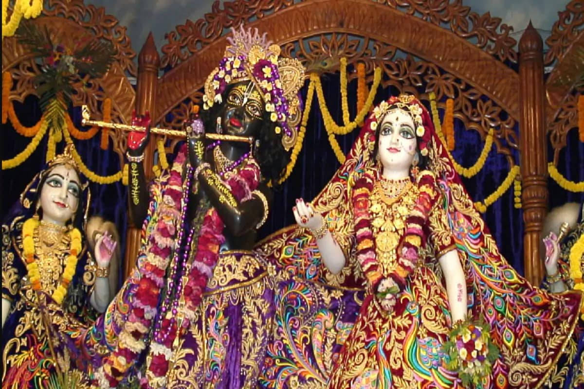Krishna Flute 桌布 壁紙 — 三位裝飾華麗的印度教神祇，身著鮮豔的金色和紫色服飾，站在裝飾著黃色花環的華麗木製天篷下。