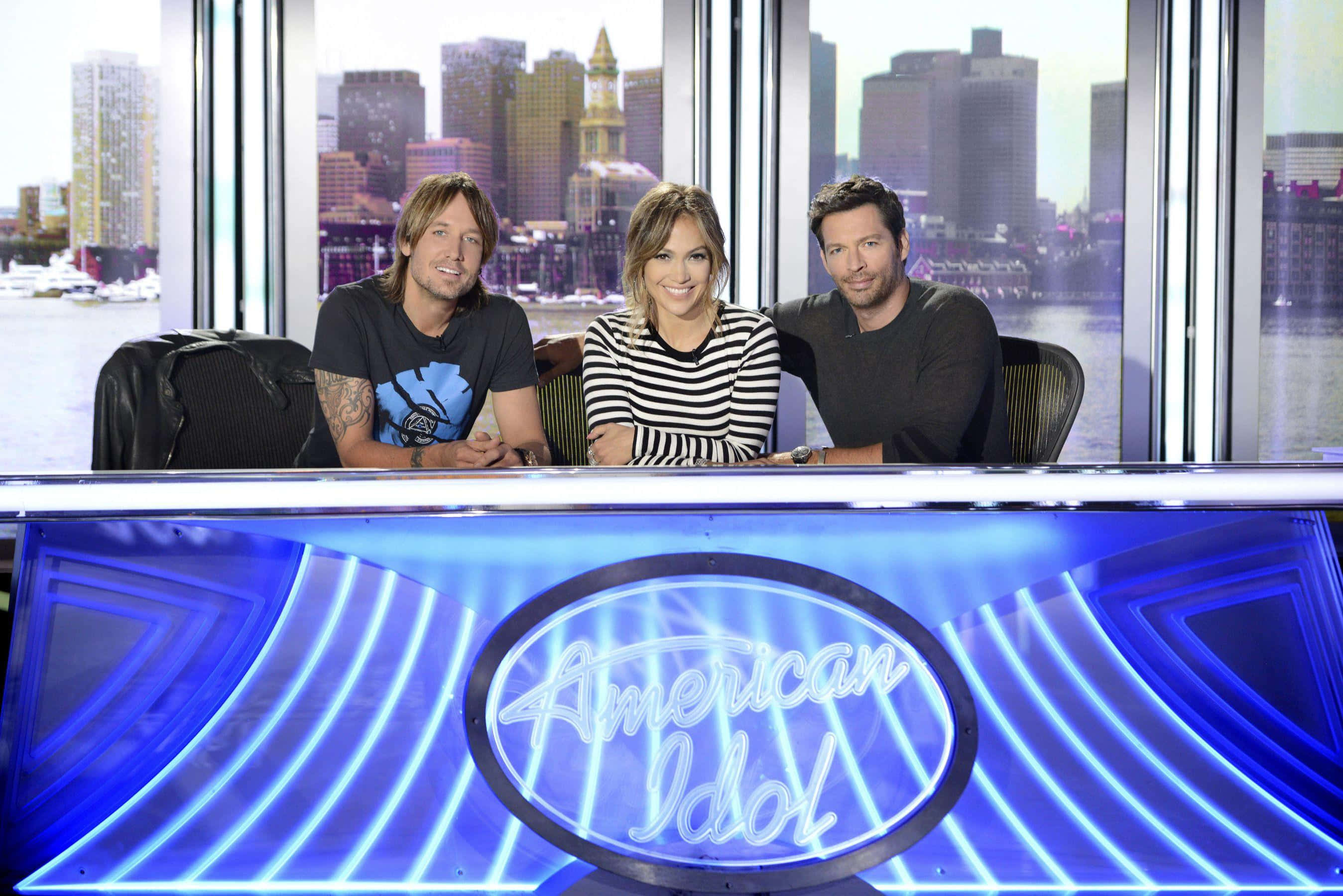 american idol 桌布 壁紙 — 三位評判坐在發光的藍色American Idol桌子後面，透過身後的大窗戶可以看到城市天際線。