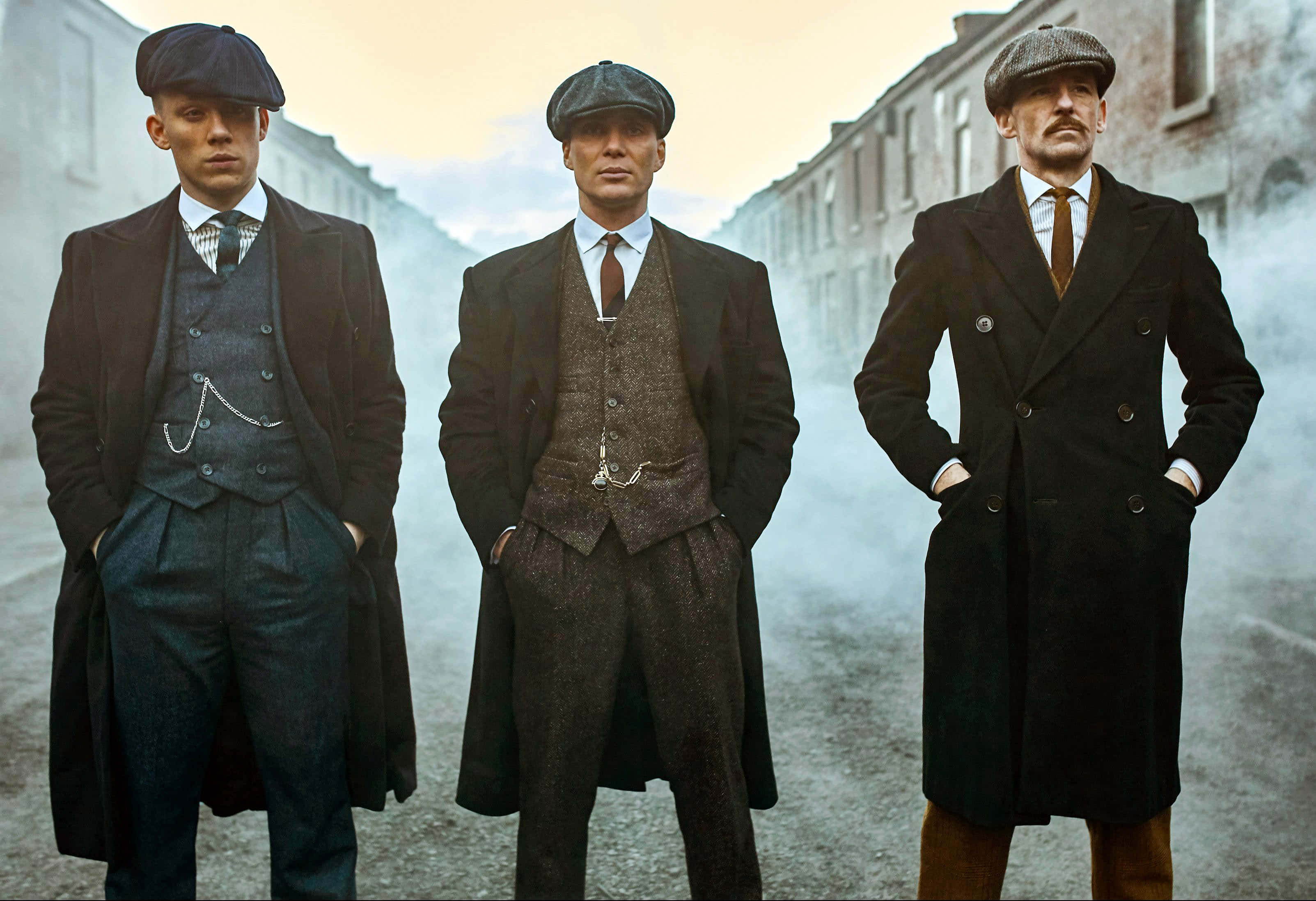 Peaky Blinders 桌布 壁紙 — 三位身穿復古西裝和扁平帽的男子，在霧氣瀰漫的城市街道上莊嚴地站立，背景是磚造建築。