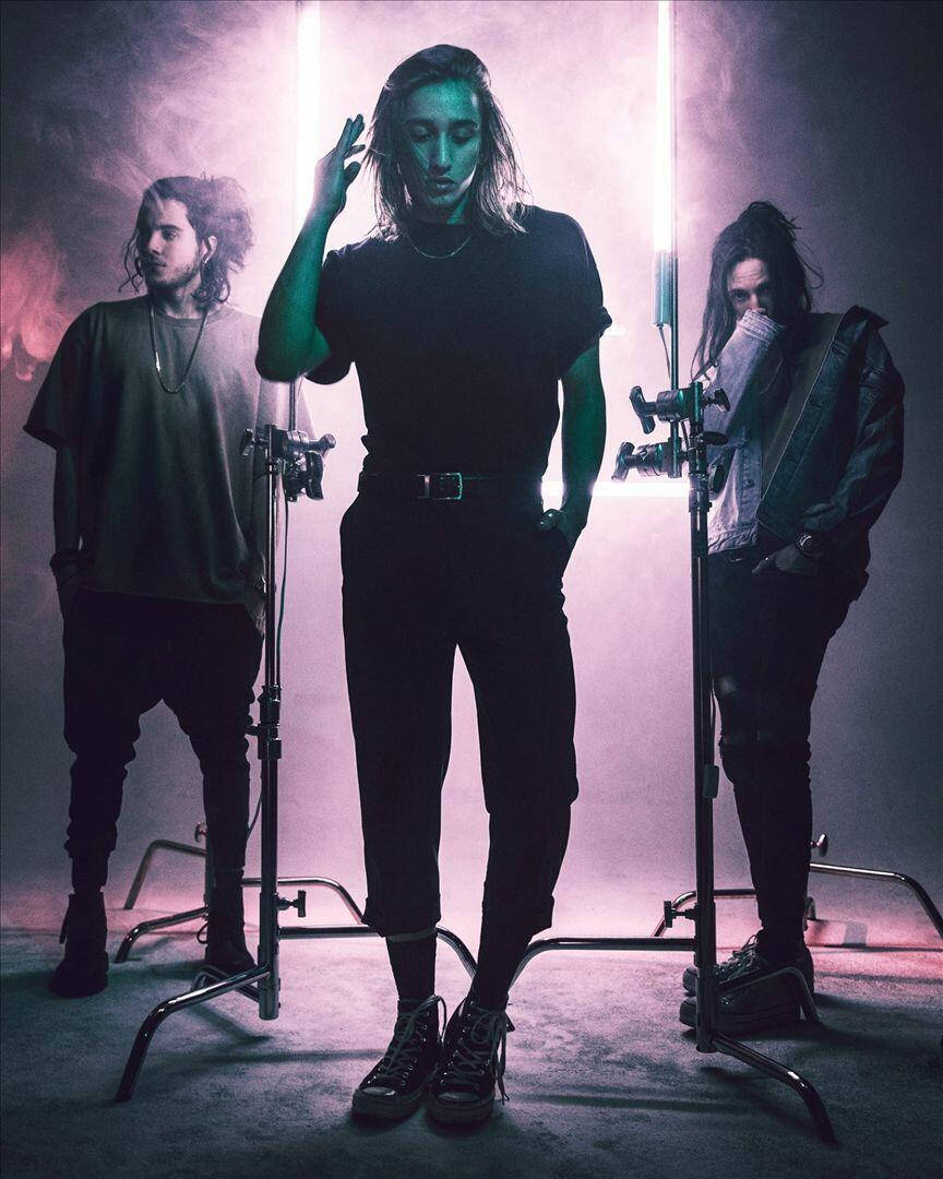 Chase Atlantic 壁紙 — 三位身穿深色衣服的音樂家站在一個煙霧瀰漫的錄音室中，被垂直的霓虹燈照亮，投射出紫色和綠色的色調。
