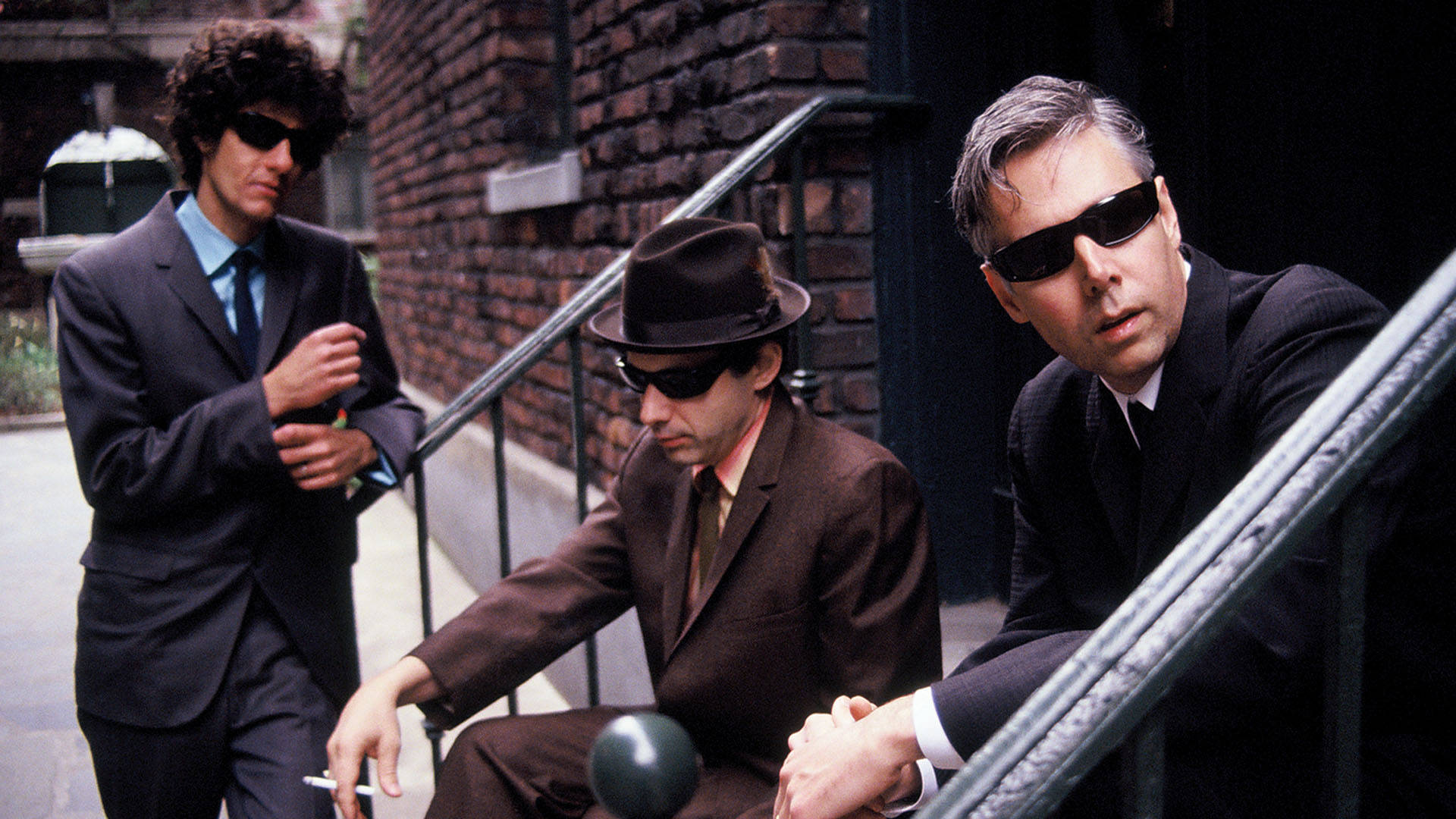 Beastie Boys《Fight For Your Right》 壁紙 — 三位身穿西裝和太陽眼鏡的男士在磚樓梯上擺姿勢，帶有復古都市氛圍。