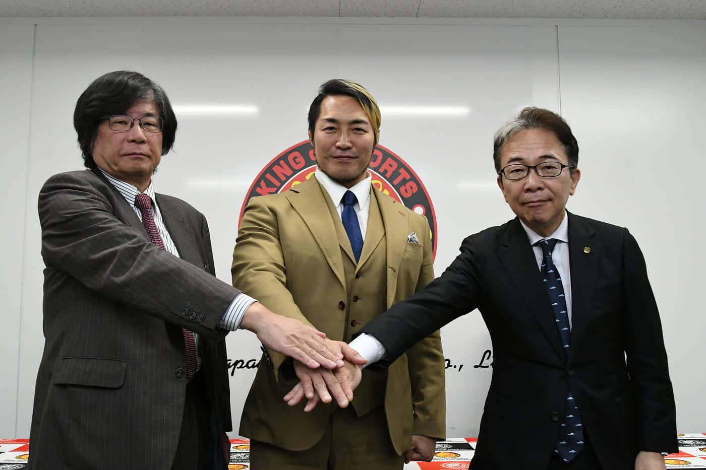  壁紙 — 三位身穿西裝的商人，在帶有紅色圓形標誌的企業牆前，將手疊在一起以示團結。