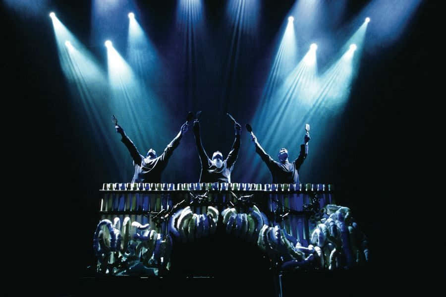 Blue Man Group 桌布 壁紙 — 三位音樂家在戲劇性的藍色舞台聚光燈下，勝利地將手臂高舉於顫音琴之上。