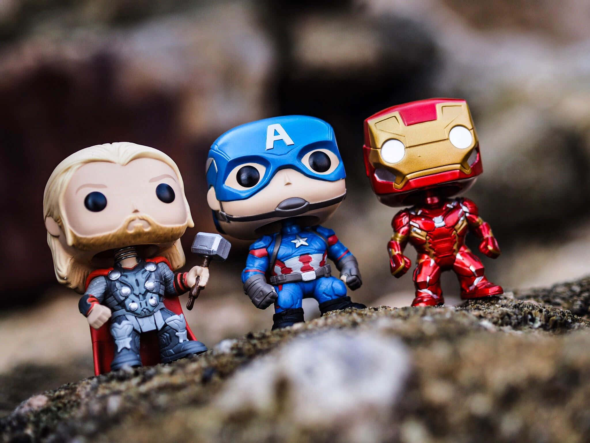 Iron Man Pop Figures 桌布 壁紙 — 三個 Funko Pop! Marvel 超級英雄 Thor、Captain America 和 Iron Man 站在岩石表面上，背景是模糊的自然景色。
