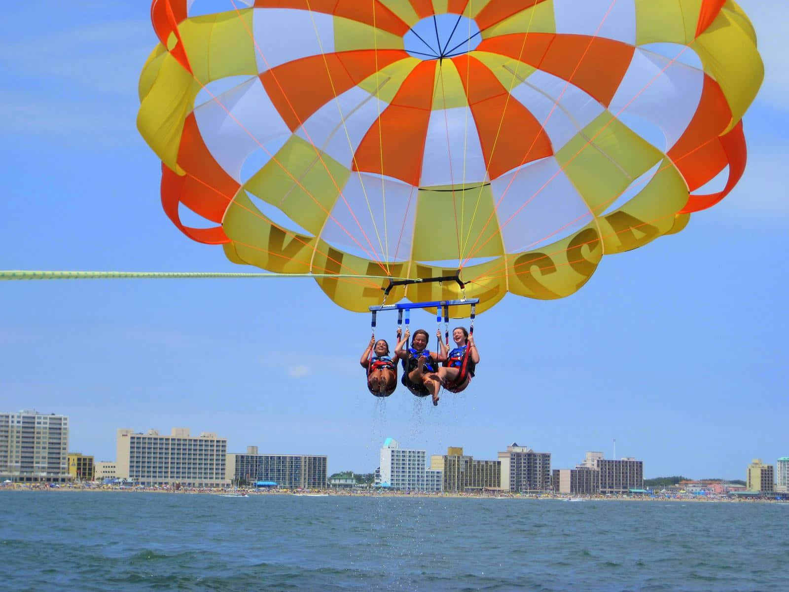 Beach Water Sports 桌布 壁紙 — 三個人喺鮮豔嘅橙色、黃色同白色降落傘上，喺碧綠海水上空快樂咁飛翔，背景係沿海城市嘅天際線。