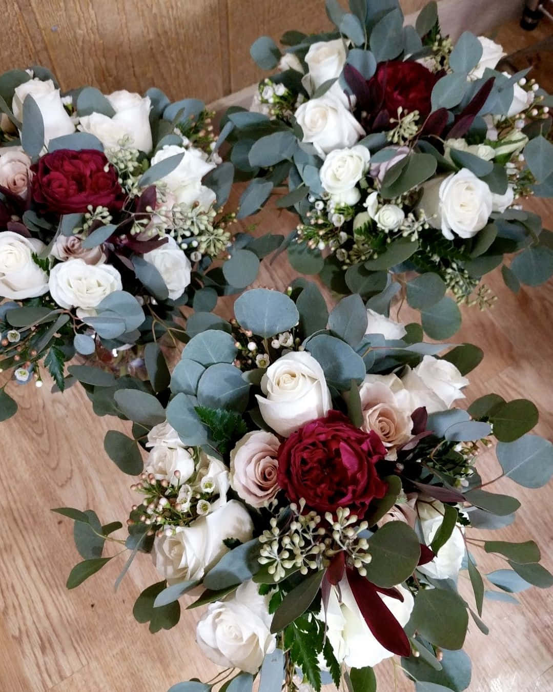Roses Wedding 桌布 壁紙 — 三個優雅的花束，包含白玫瑰、酒紅色牡丹和尤加利葉，擺放在木地板上。