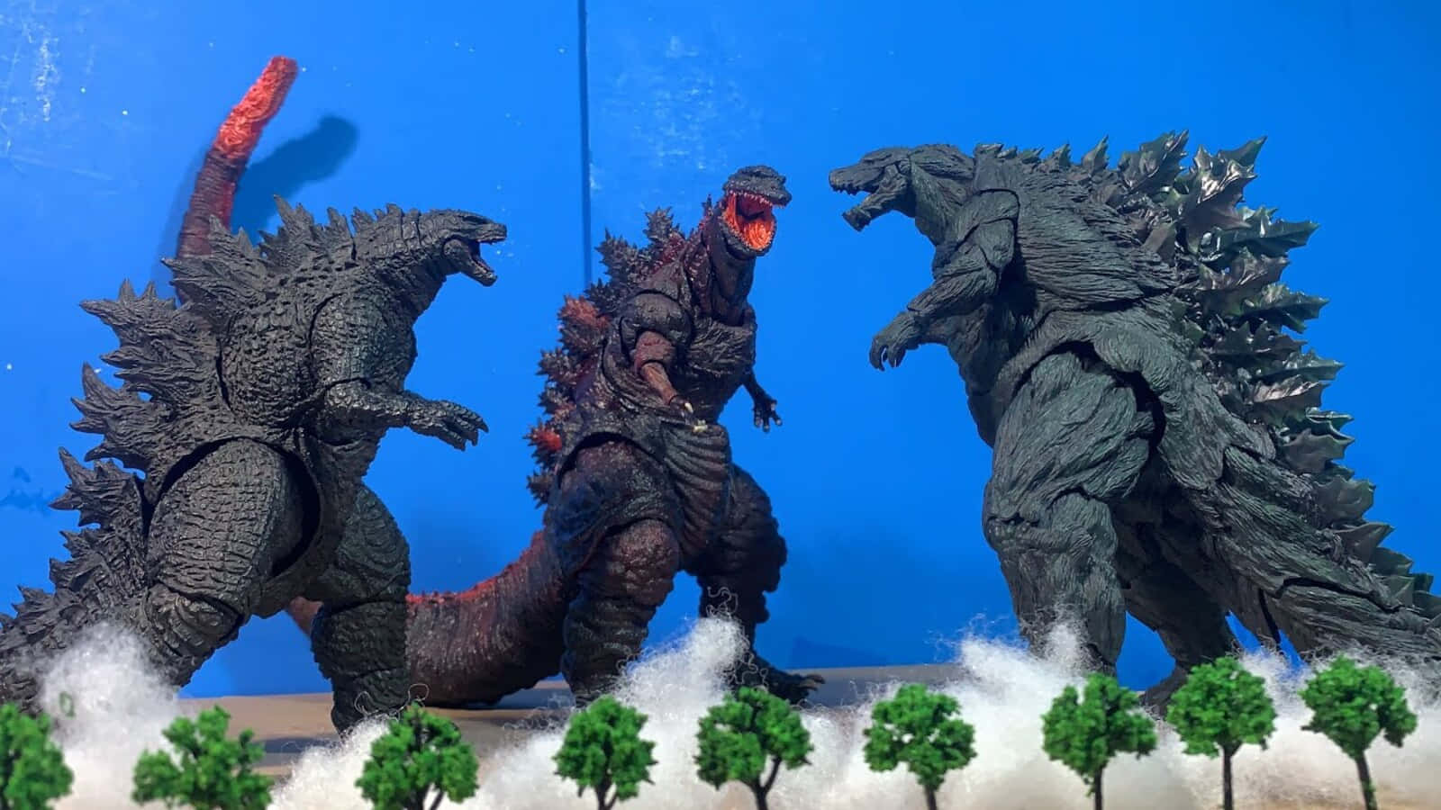 Godzilla Earth 桌布 壁紙 — 三個哥斯拉動作人偶以戰鬥姿勢站立在雪地地形上，配有微型樹木，背景為純藍色。