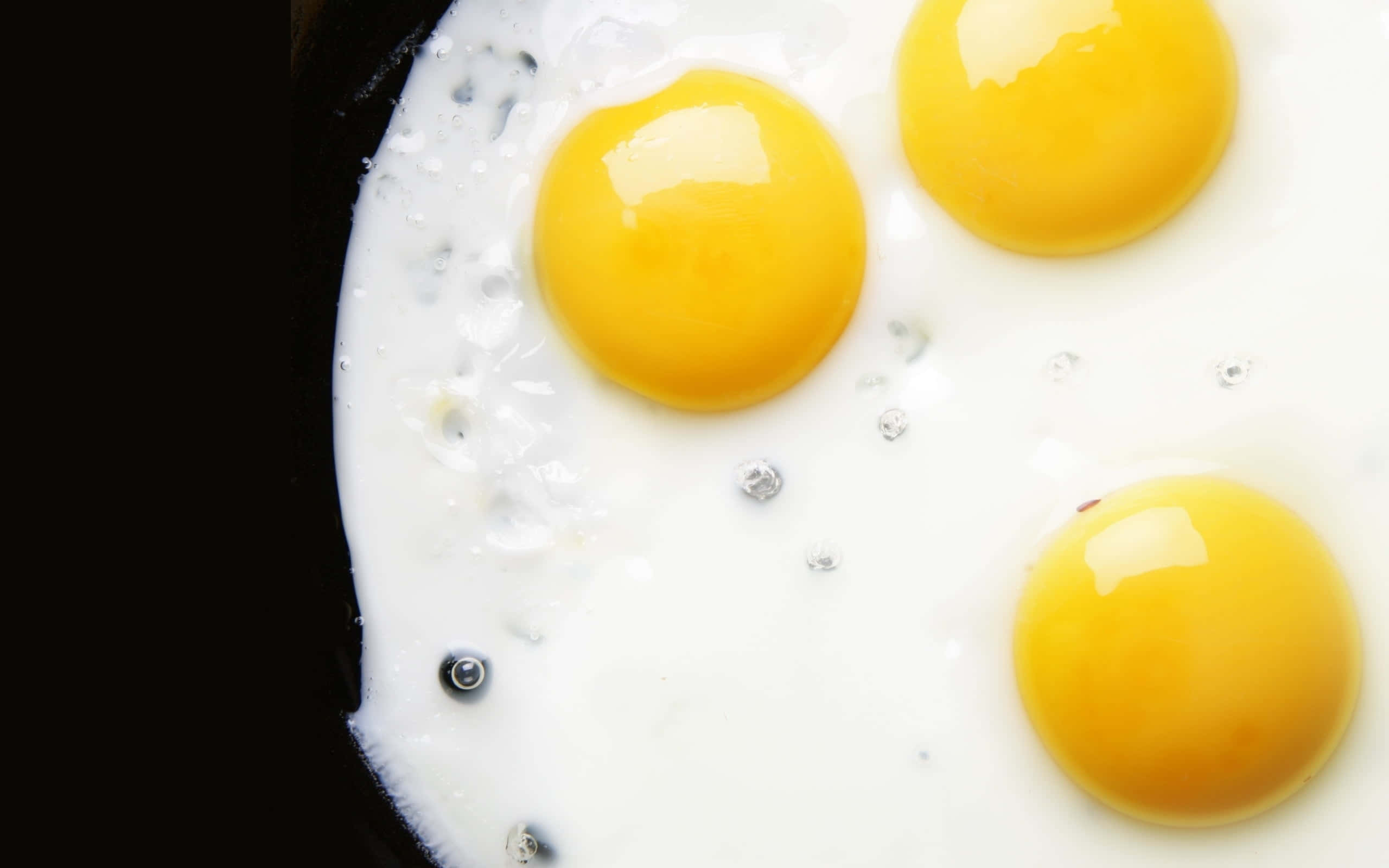 Egg White 桌布 壁紙 — 三個完美圓形的太陽蛋在黑色平底鍋中滋滋作響，蛋白冒泡，蛋黃閃亮。