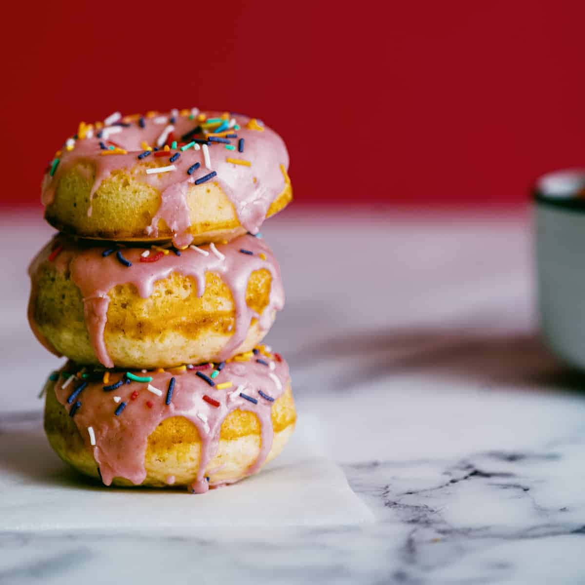 Glazed Donut 桌布 壁紙 — 三個帶有彩色糖粒的釉面冬甩堆疊在大理石表面上，背景為大膽的紅色。