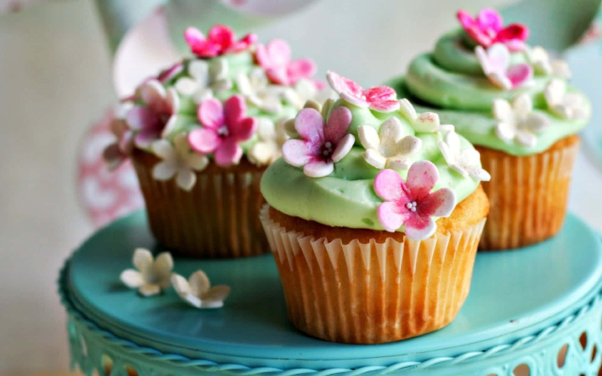 Spring Desserts 桌布 壁紙 — 三個帶有淺綠色糖霜和粉白色糖花的杯子蛋糕，放在一個藍綠色的蛋糕架上。