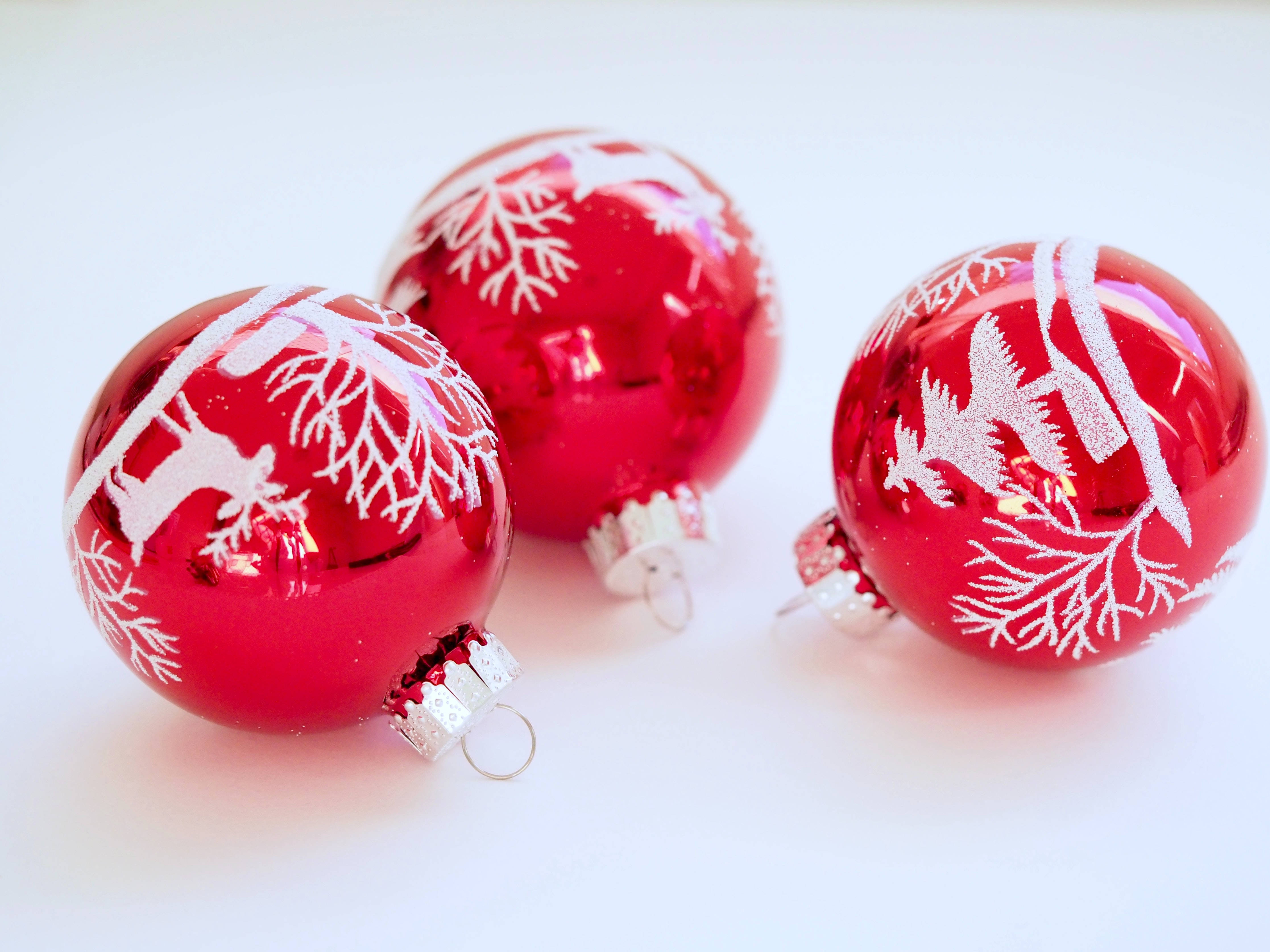 Christmas Balls 桌布 壁紙 — 三個帶有白色雪花和馴鹿圖案的亮面紅色聖誕裝飾品，放置在明亮的白色表面上。