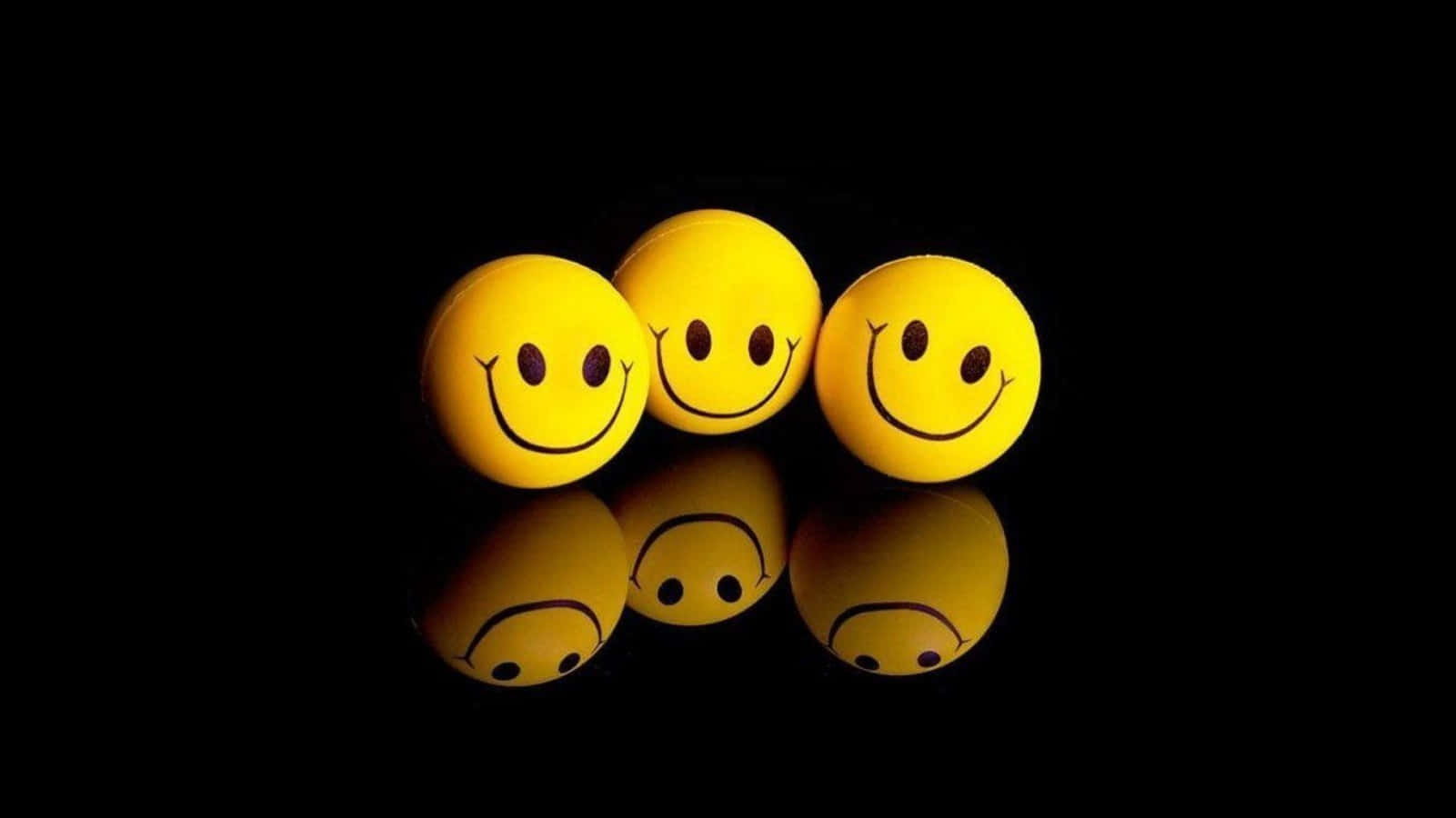Smiley 桌布 壁紙 — 三個帶有簡單黑色面孔的亮黃色笑臉球放置在純黑色背景下的反光表面上。