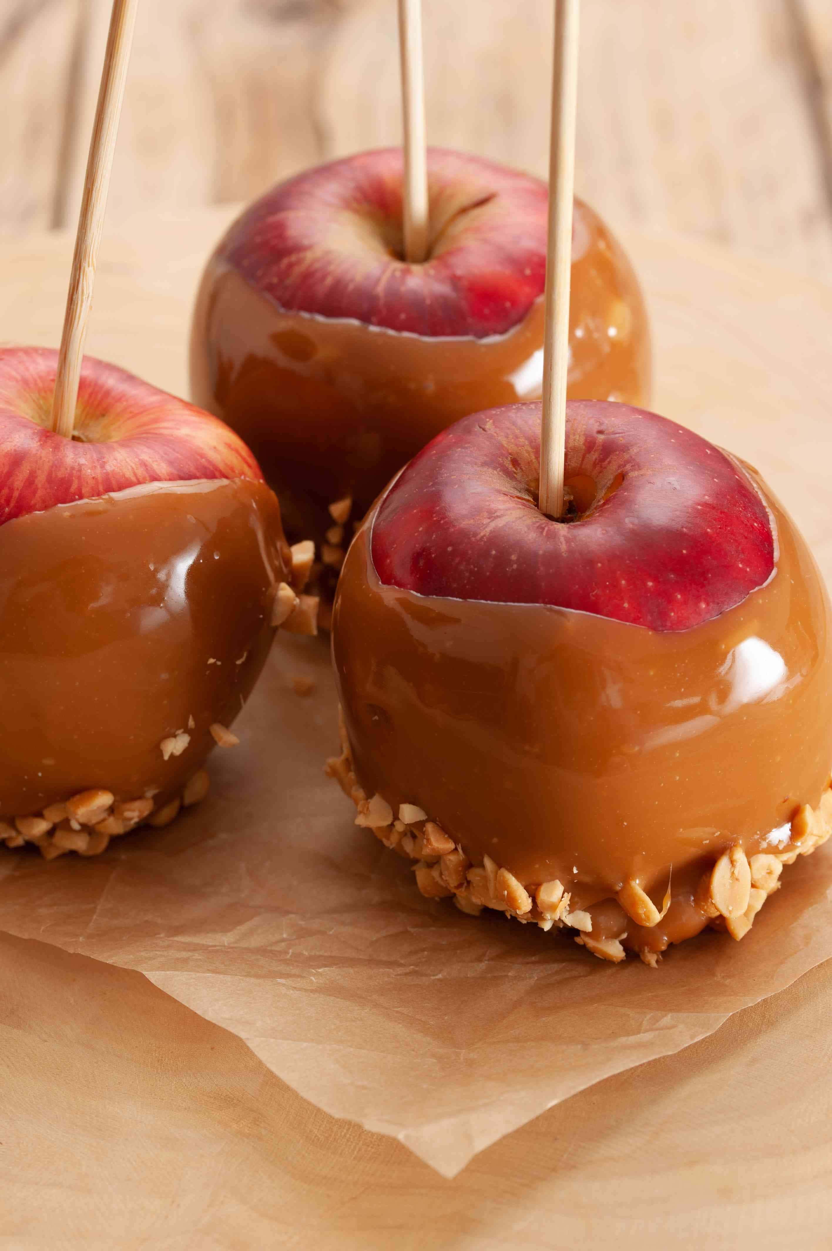 Caramel Apples 桌布 壁紙 — 三個帶有花生配料的光澤焦糖蘋果放在木質表面上的棕色牛油紙上。