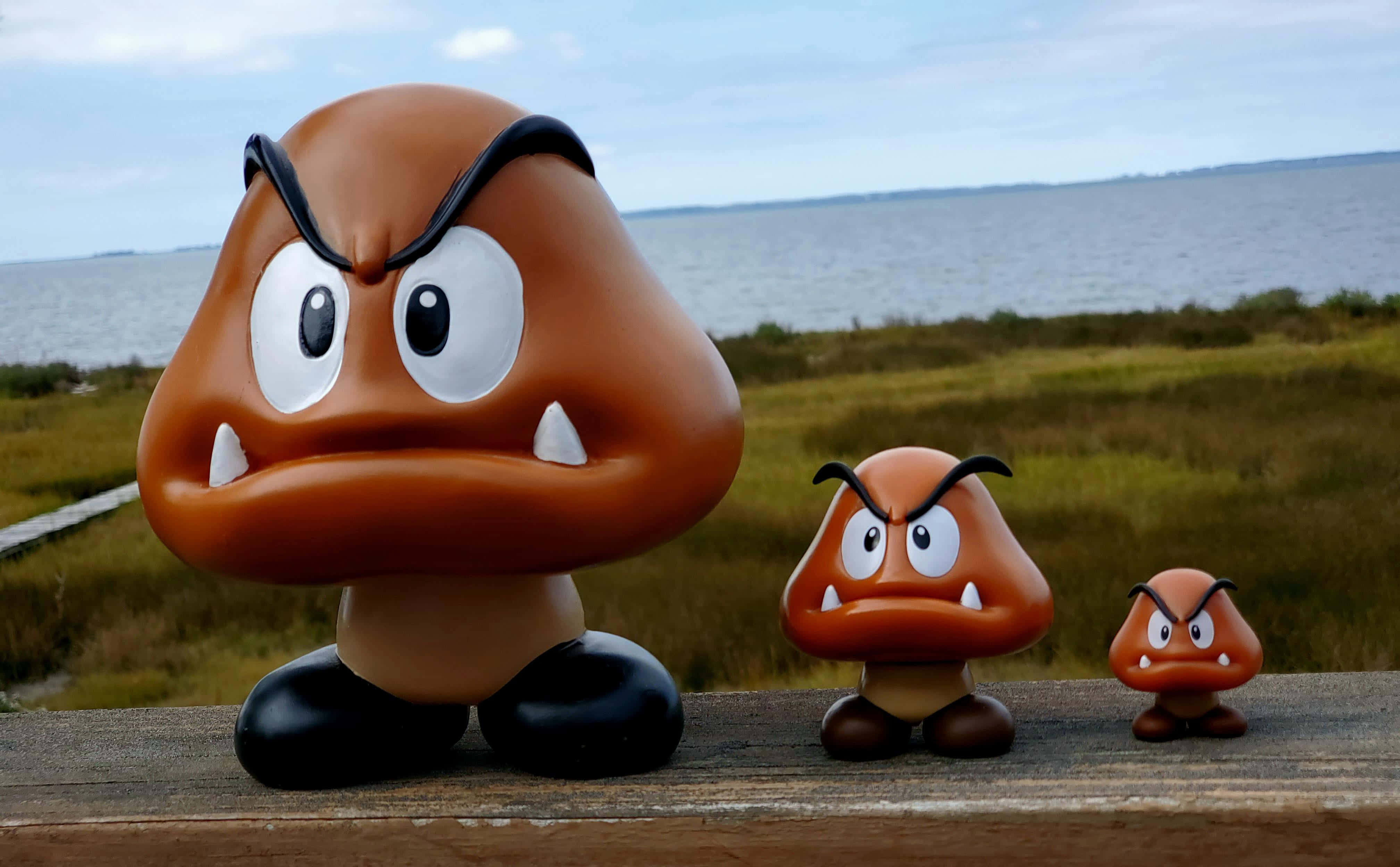 Goomba 桌布 壁紙 — 三個憤怒嘅Goomba公仔企喺木欄杆上一字排開，俯瞰住一片草地沼澤同平靜嘅水面，天空密雲。