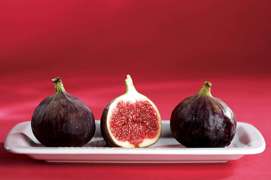 Purple Figs 桌布 壁紙 — 三個成熟的無花果，其中一個切開露出多汁的紅色果肉，放在白色碟子上，背景為鮮紅色。
