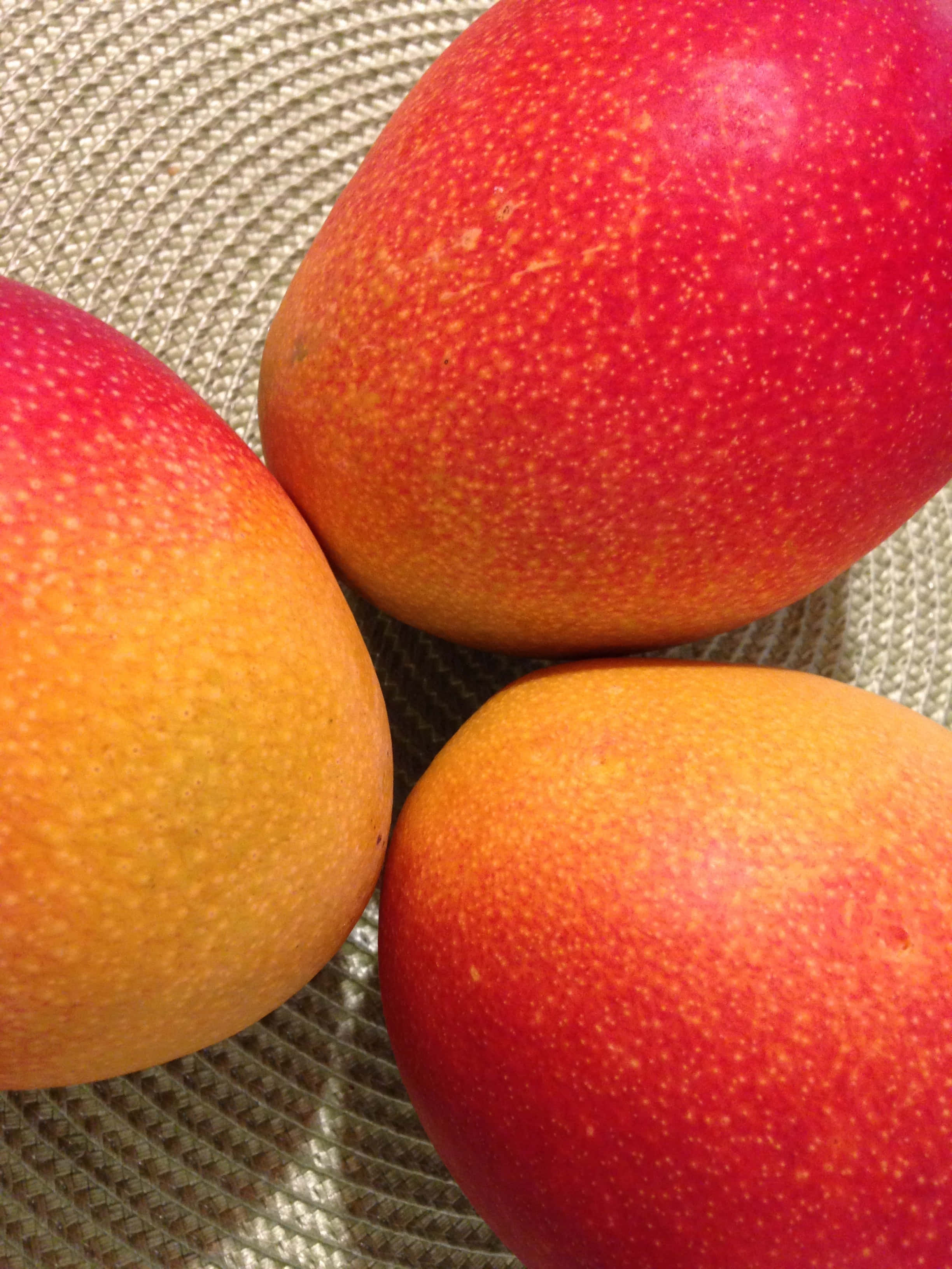 Mango 桌布 壁紙 — 三個成熟的芒果，帶有紅色和橙色的果皮，放在編織紋理的表面上。