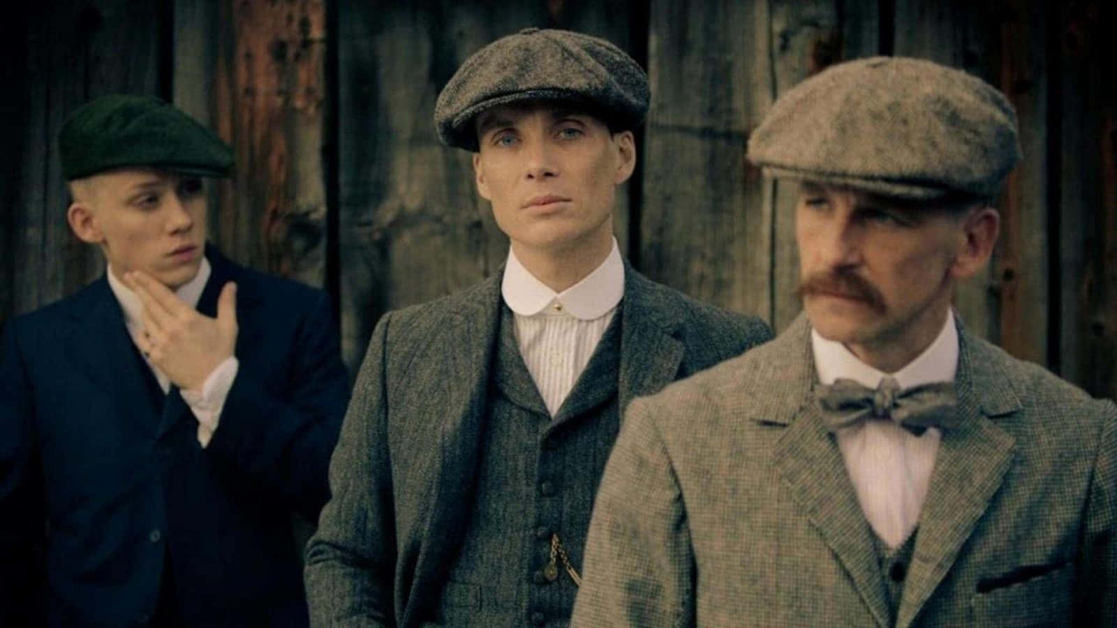 Peaky Blinders 桌布 壁紙 — 三個戴著復古平頂帽、身穿西裝的男人，莊嚴地站在一面風化的木牆前。